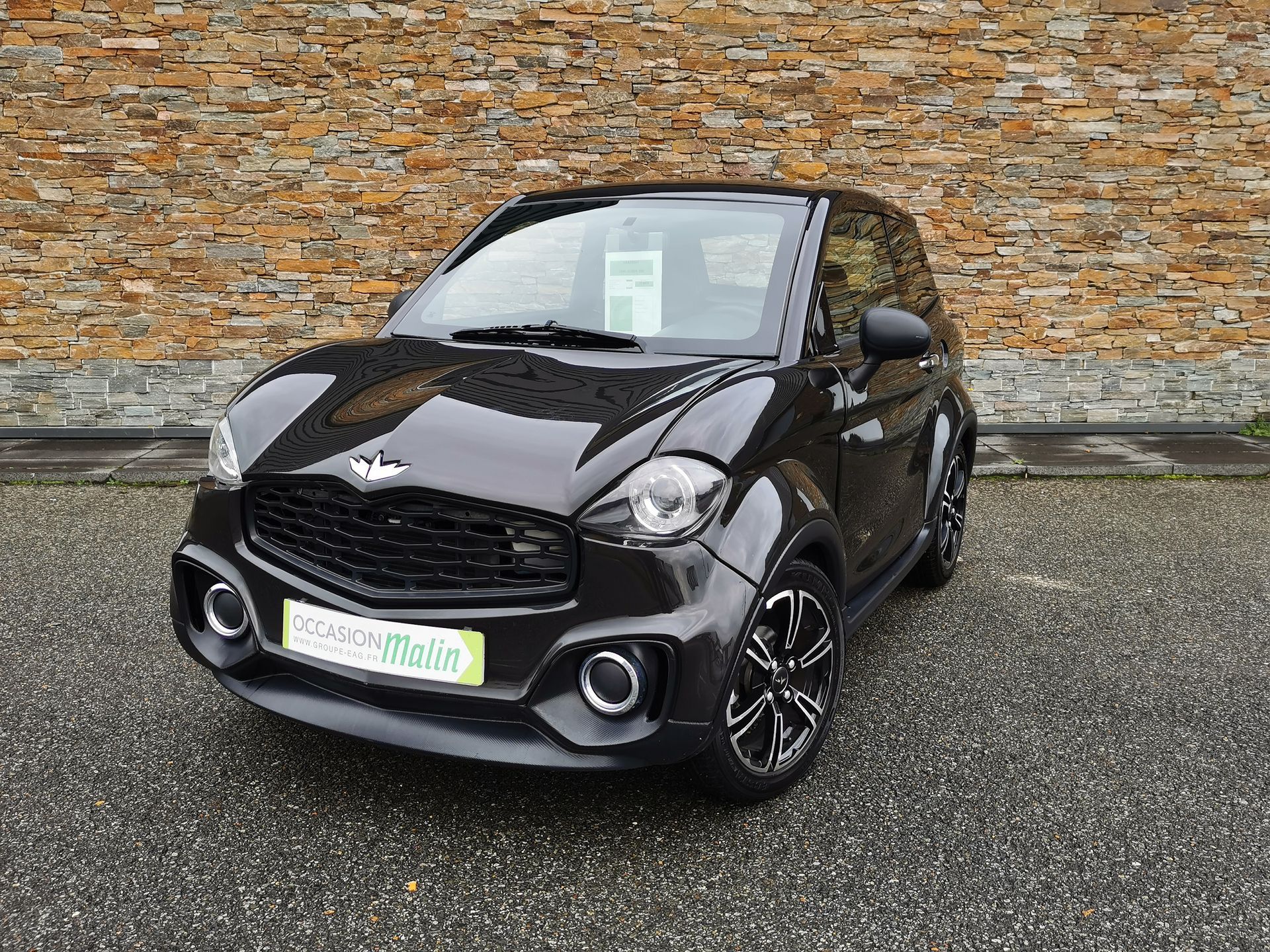 Voiture électrique Aixam noire garée devant un mur de briques.