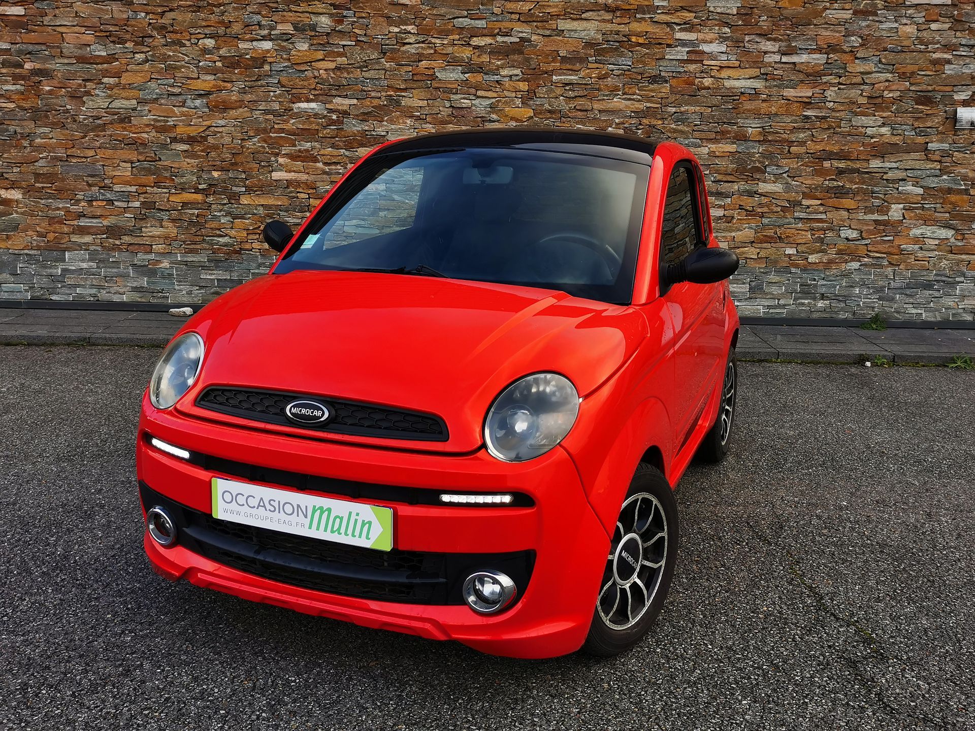 Voiture compacte rouge garée devant un mur de briques, toit noir, petites roues.