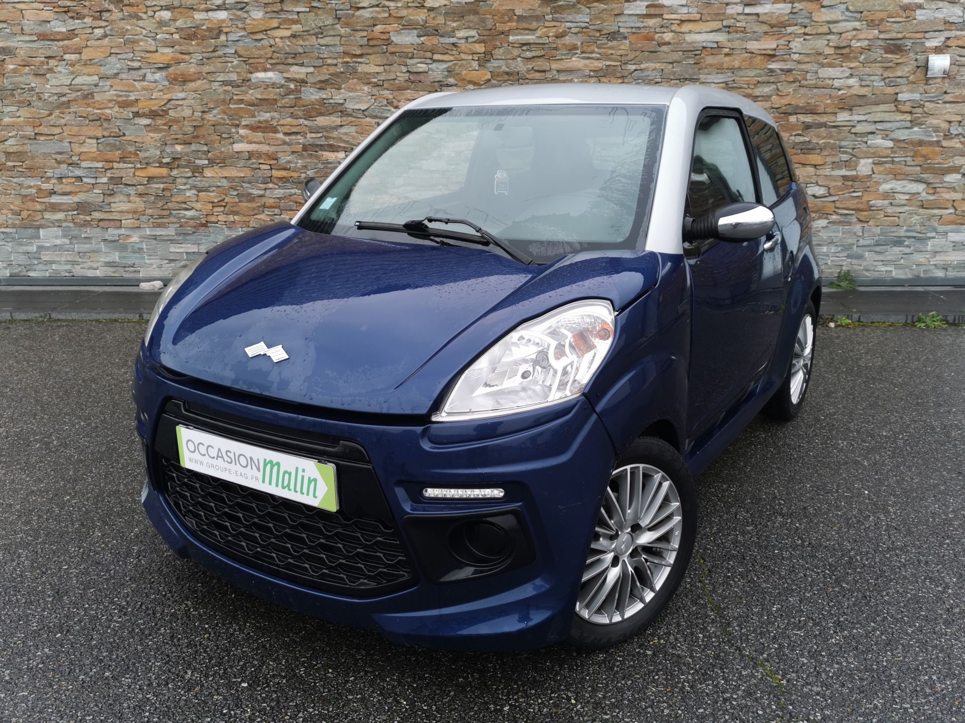 Une microvoiture deux portes bleue et argentée est garée devant un mur de briques.