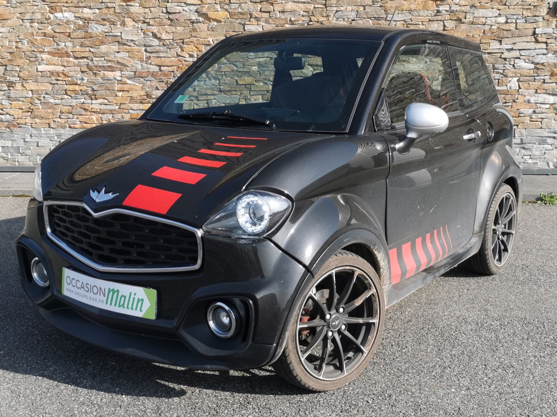 Voiture sans permis Aixam noire à bandes rouges, garé devant un mur de briques.
