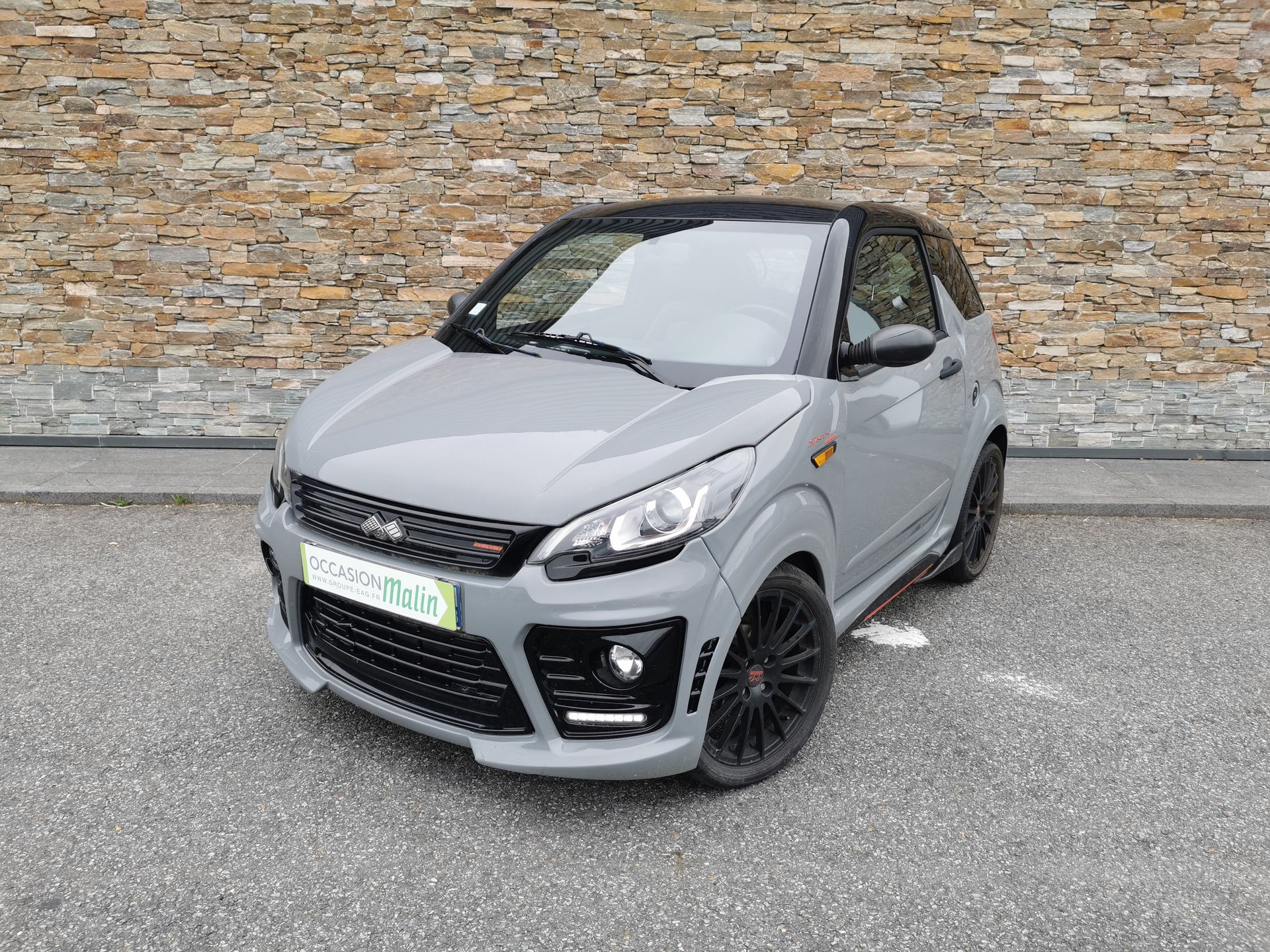 Voiture Aixam grise garée sur l'asphalte, garnitures noires, devant un mur de pierre.