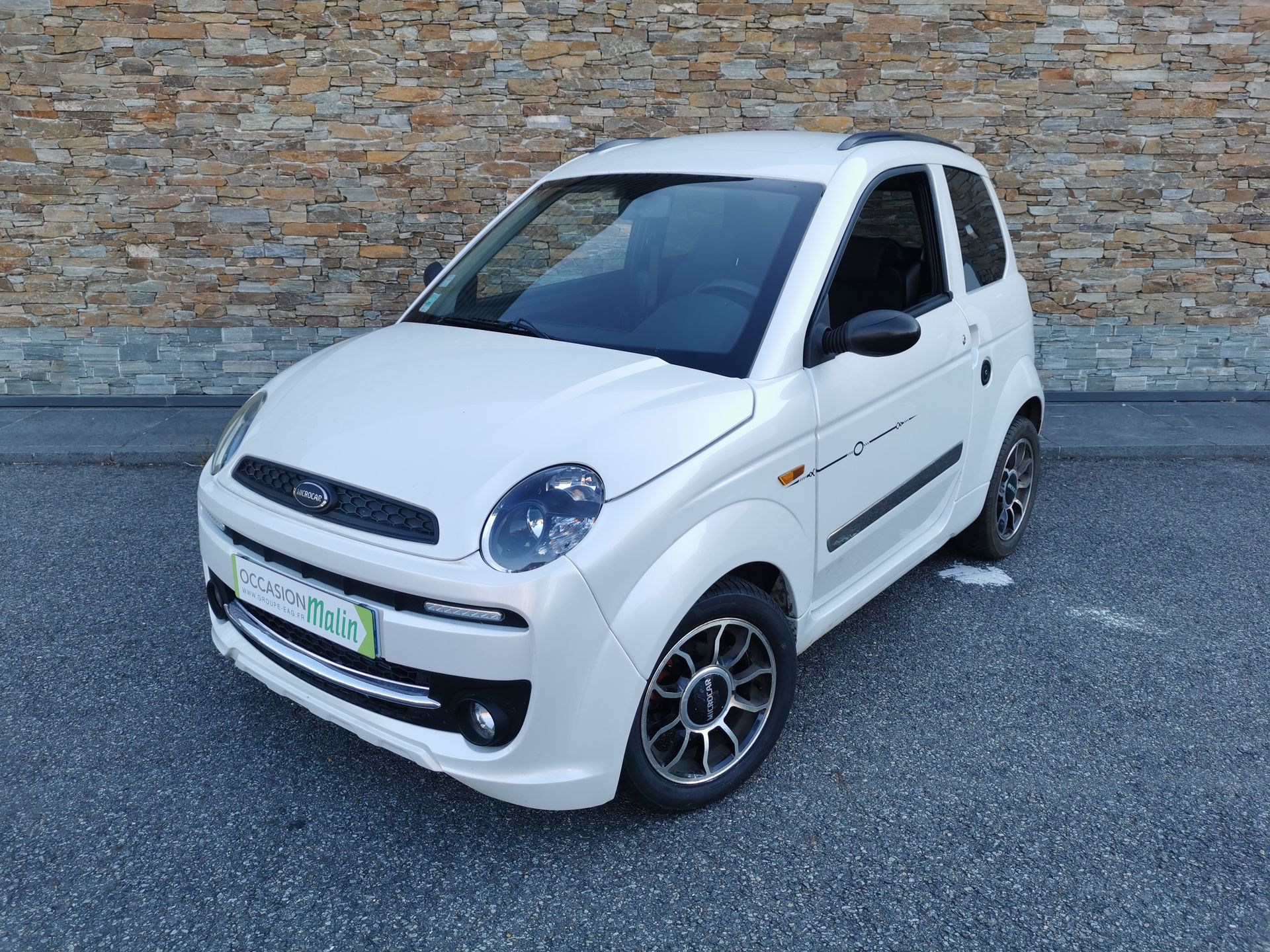 Une microvoiture blanche est garée sur le trottoir devant un mur en pierre.