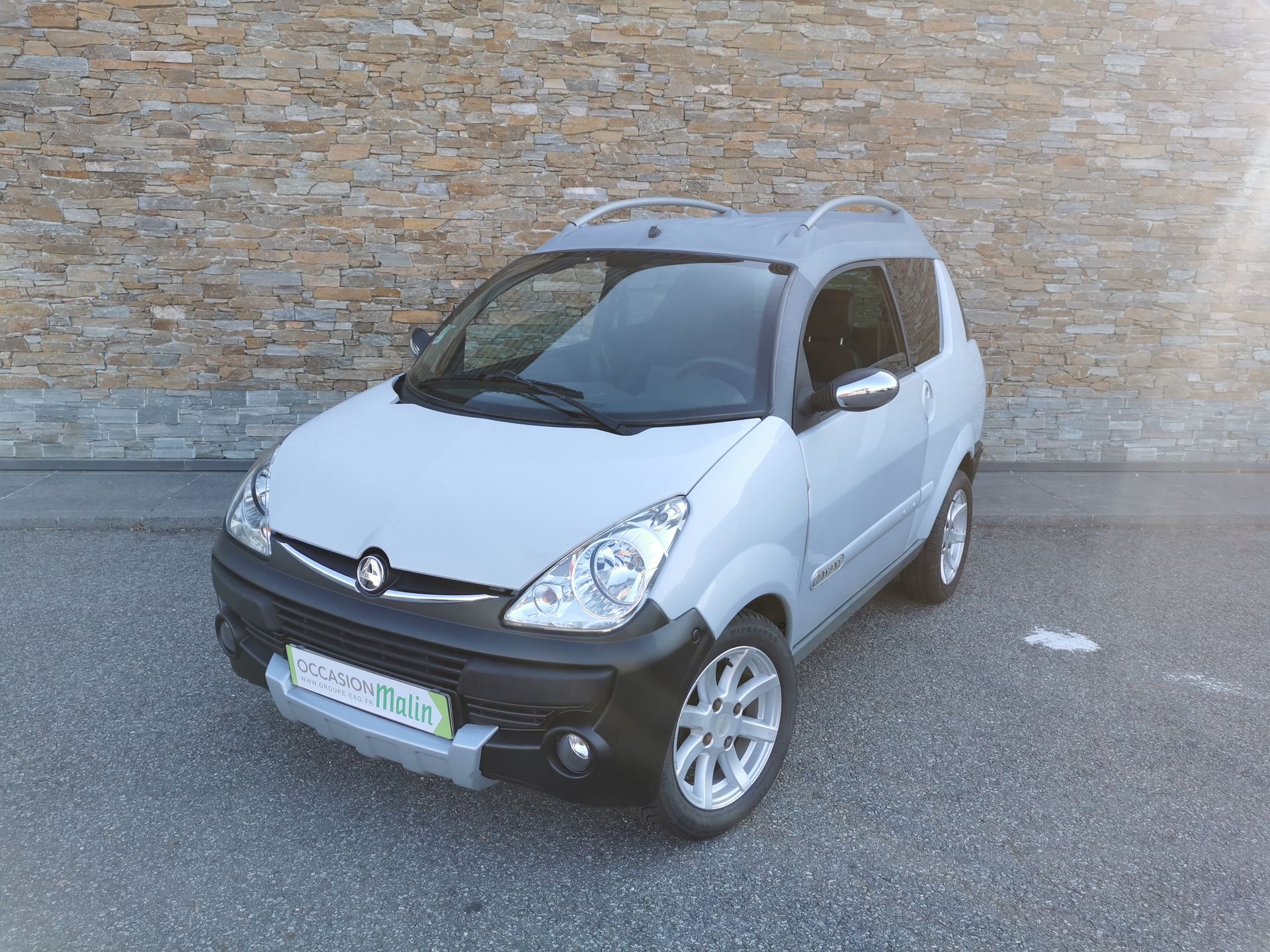 Une voiture sans permis Aixam argentée garée devant un mur en pierre.