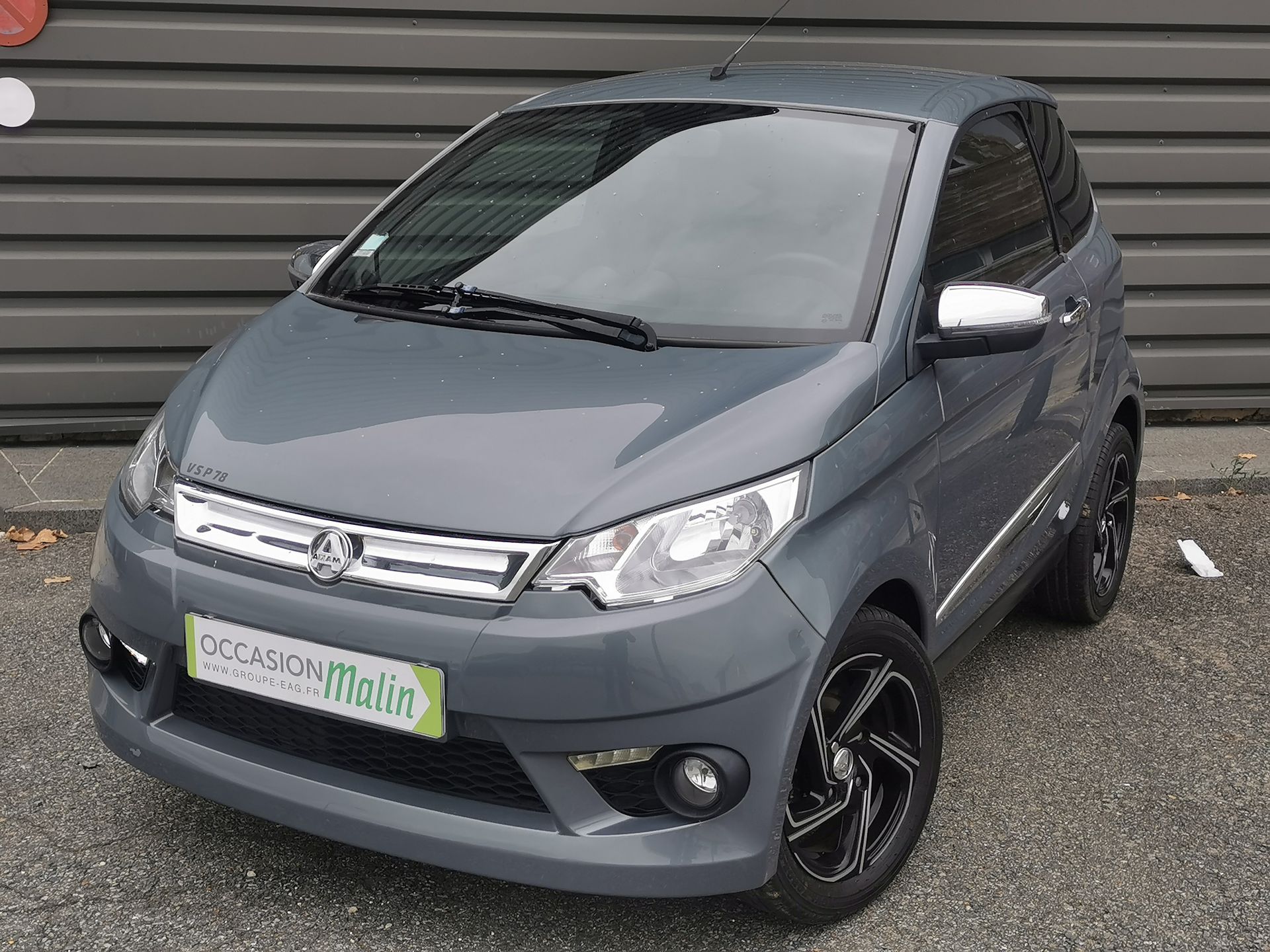 Voiture de ville grise Aixam, garée devant un bâtiment gris.