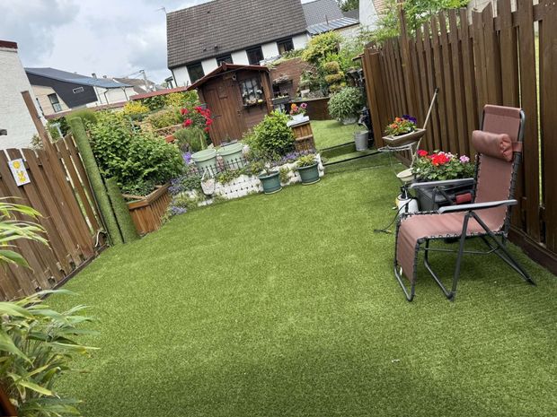 Arrière-cour verdoyante avec gazon artificiel, clôture brune, une chaise et des parterres de fleurs.