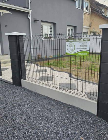 Clôture en métal noir avec allée en gravier gris et panneau devant un bâtiment gris.