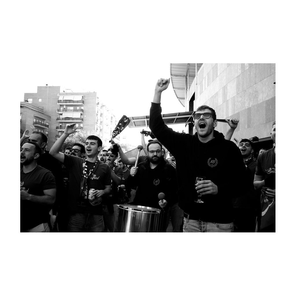 Grupo de aficionados al deporte animando, levantando los brazos, algunos con bebidas, a las afueras de un estadio.