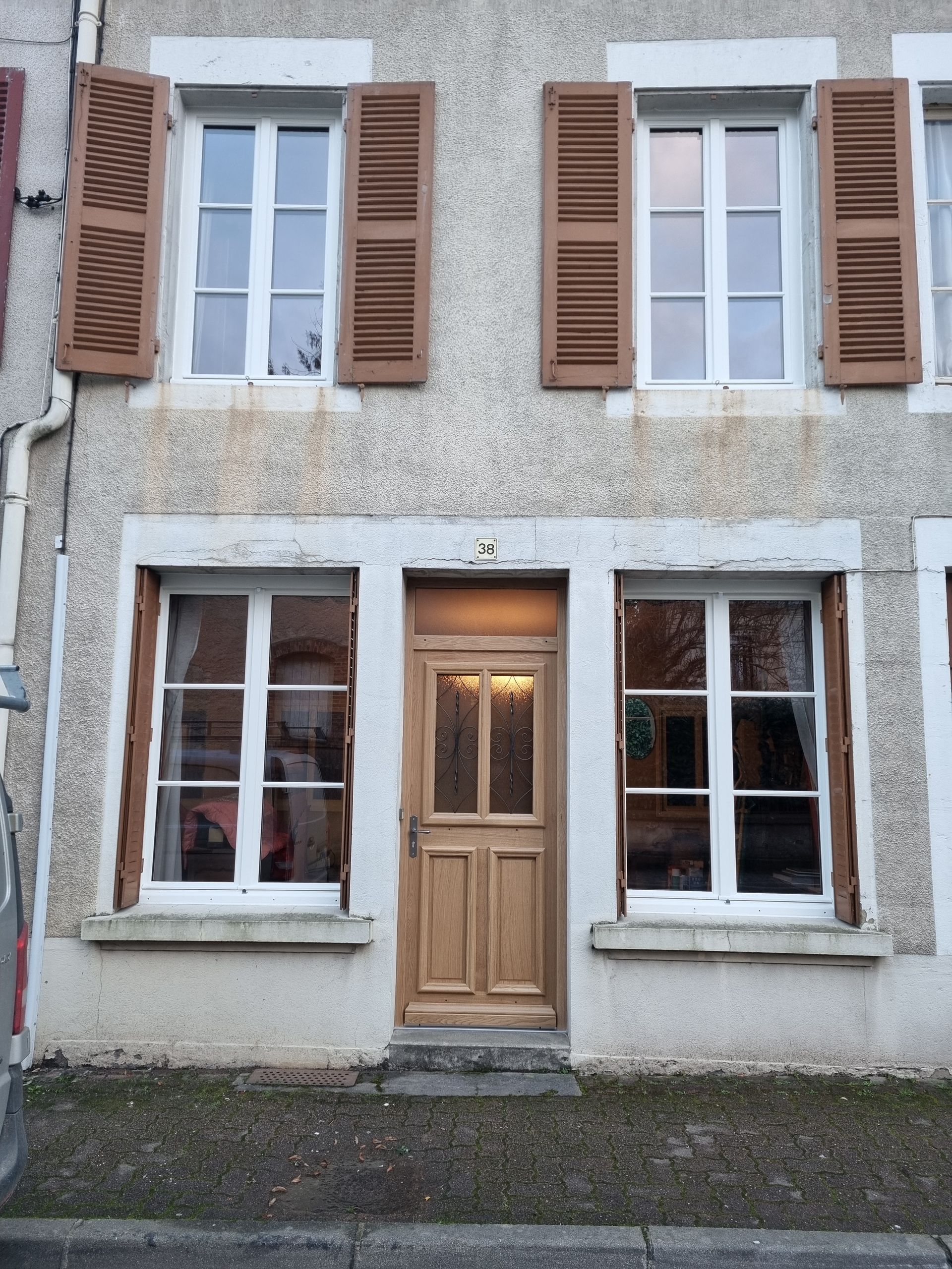 Façade d’une petite maison en pierre, avec des fenêtres blanches, des volets bruns et une porte d’entrée en bois.