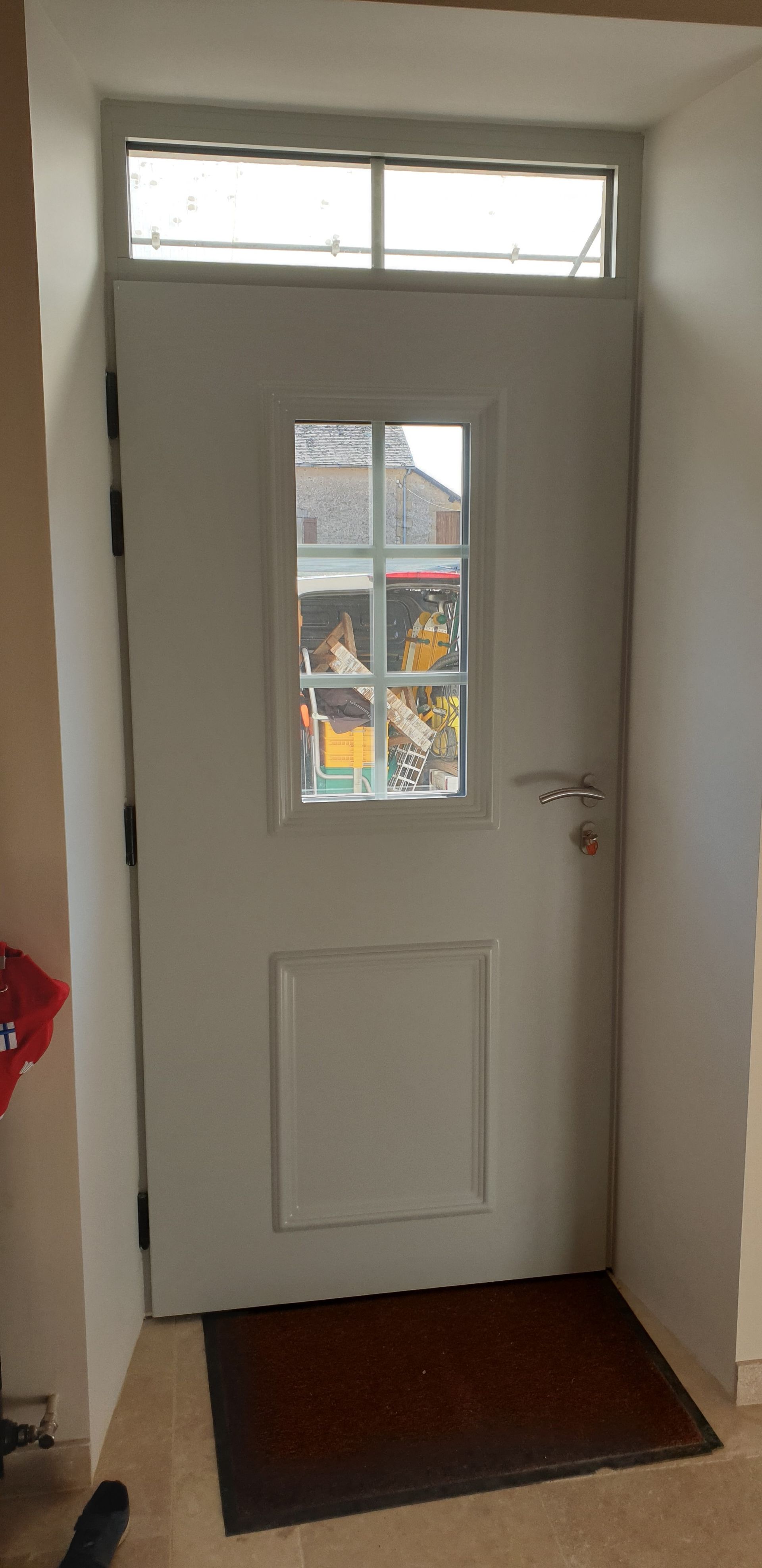 Porte d’entrée blanche avec une petite fenêtre et une imposte, vue depuis un couloir intérieur.