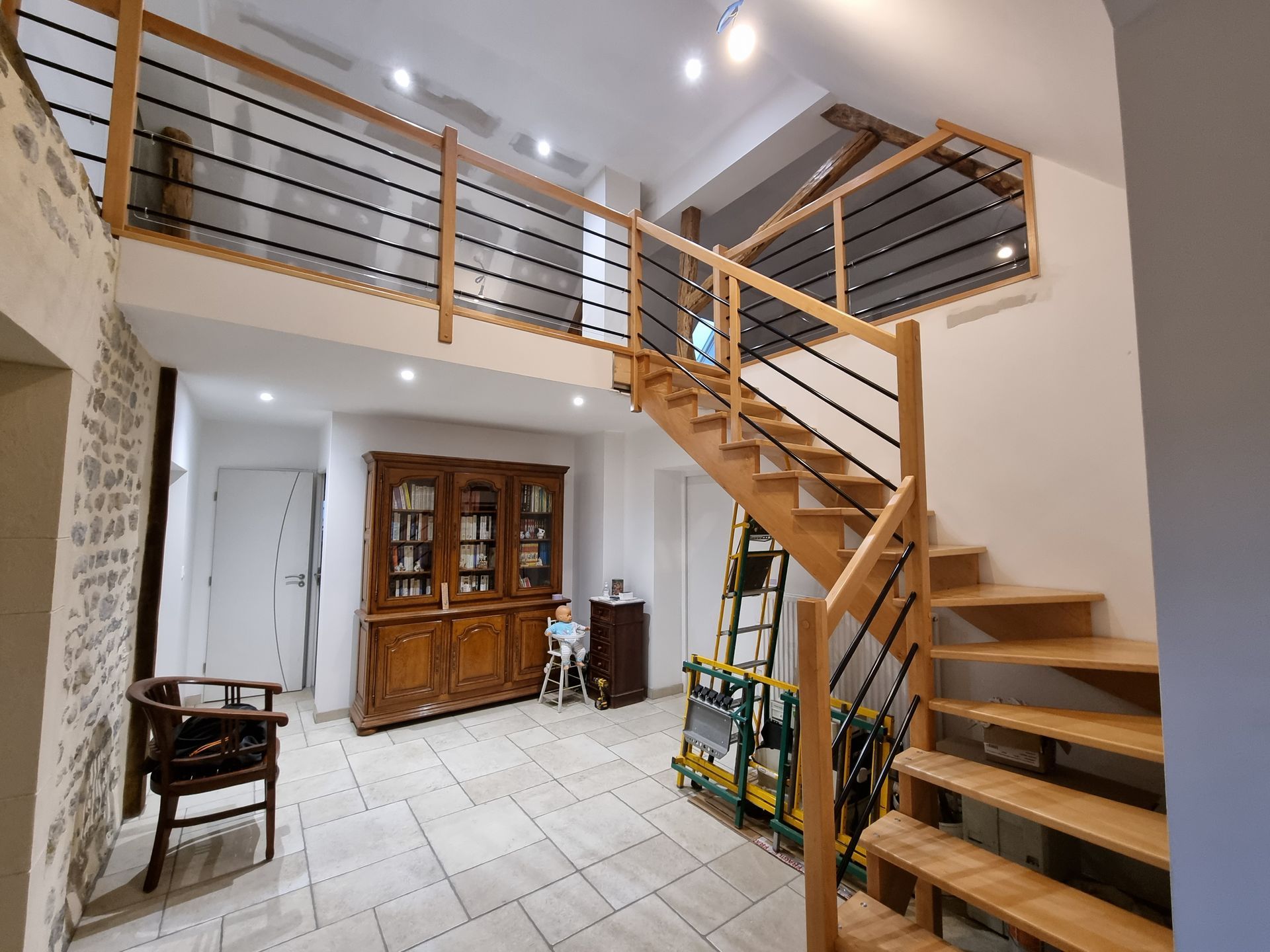 Salon moderne avec un escalier en bois, un sol carrelé et une balustrade de balcon sous des spots encastrés.