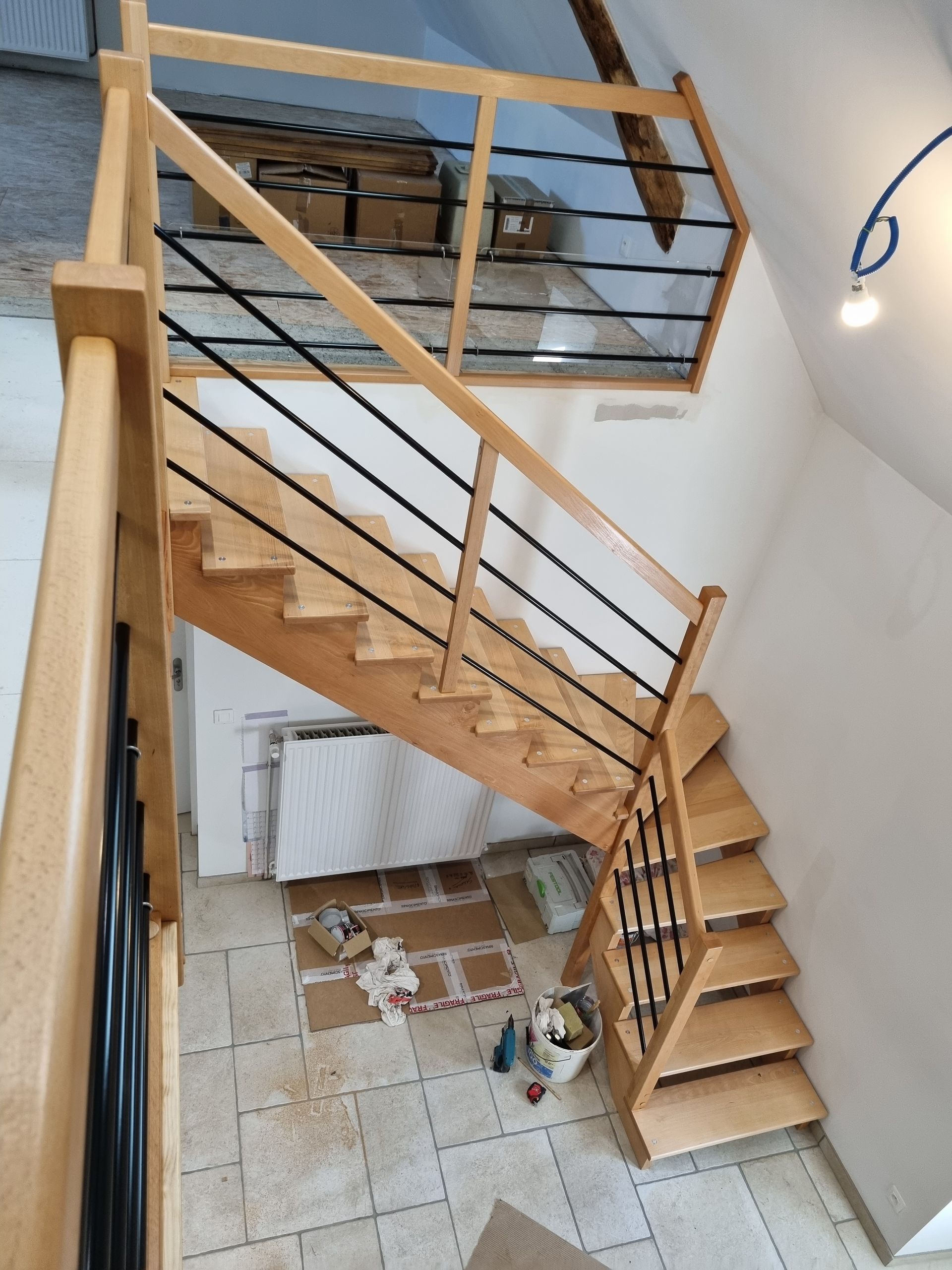 Escalier intérieur avec des marches en bois, une rampe noire et un sol carrelé dans une cage d’escalier lumineuse.