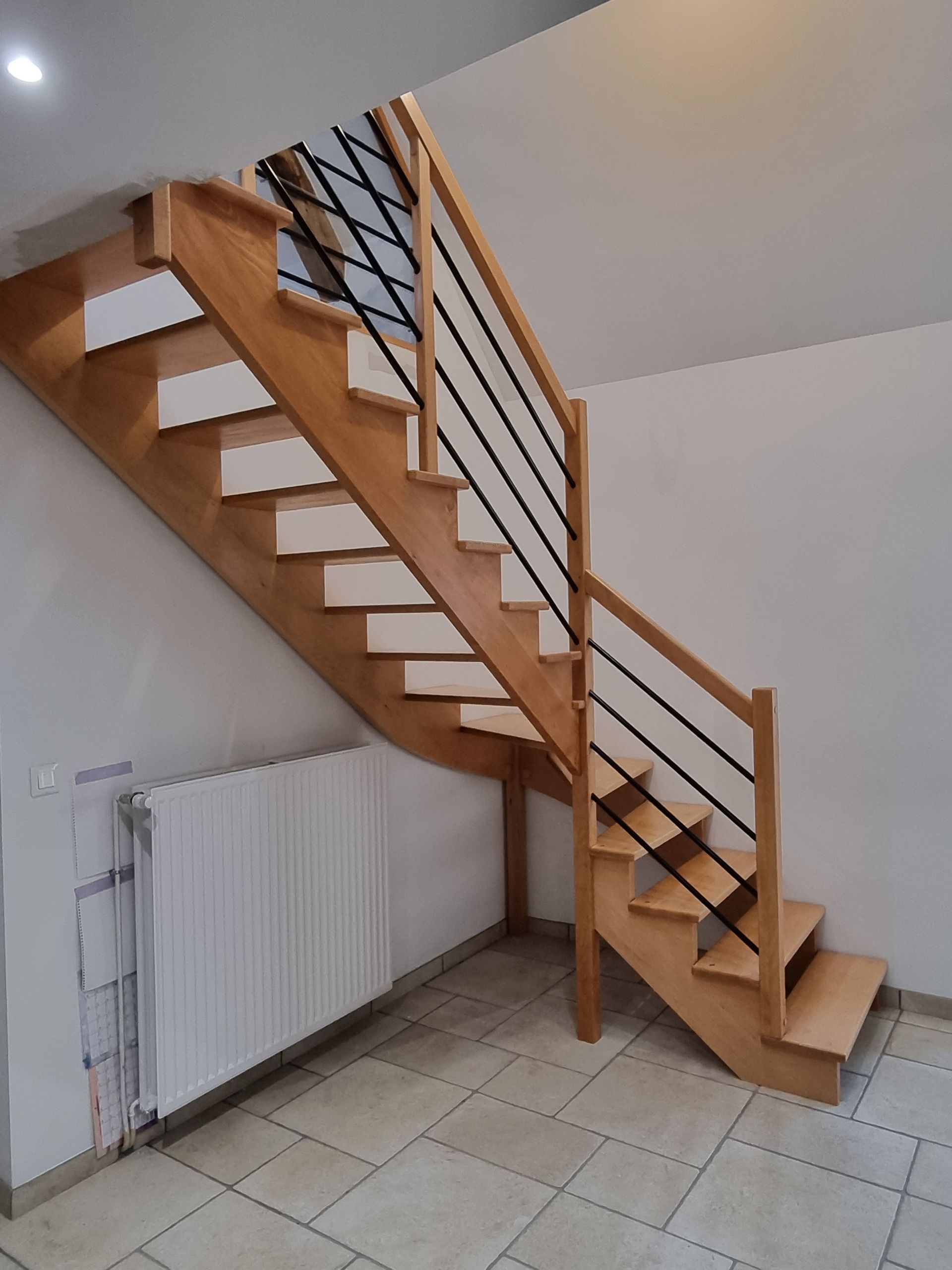 Escalier en bois avec rampe dans un intérieur carrelé, à côté d’un radiateur blanc.