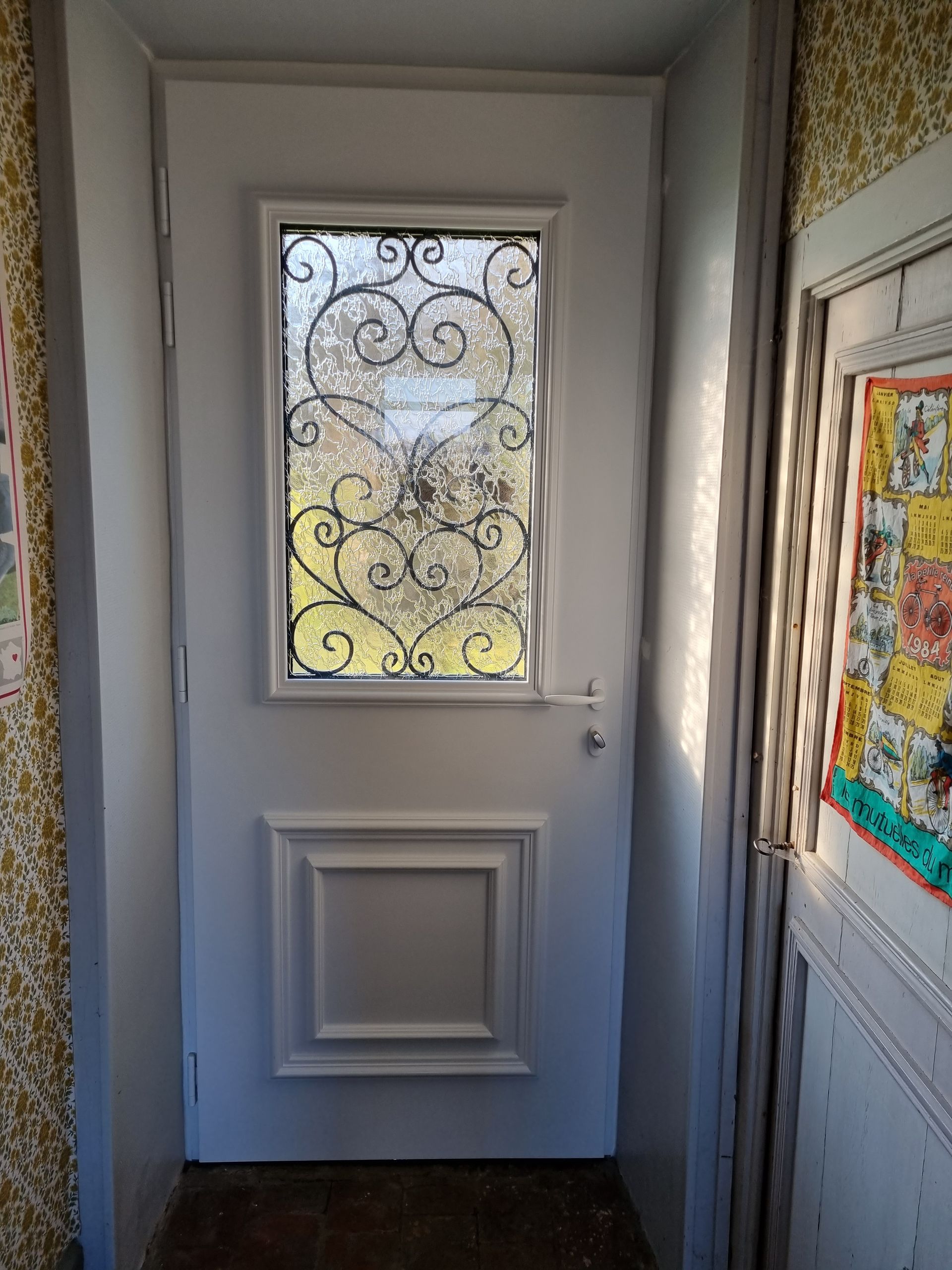 Porte d’entrée blanche avec une vitre décorative dépolie, dans un couloir étroit aux murs lambrissés et au papier peint à motifs.