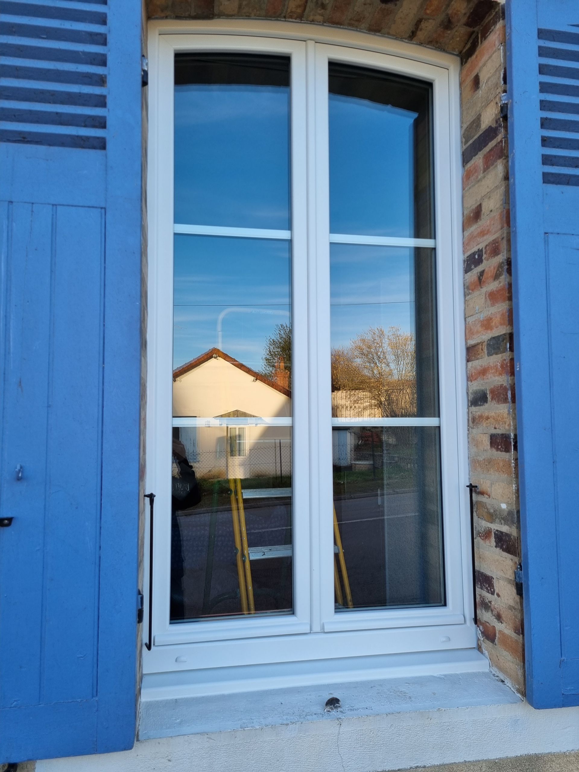 Fenêtre aux volets bleus et aux encadrements blancs, où se reflètent des maisons et des arbres dans les vitres.