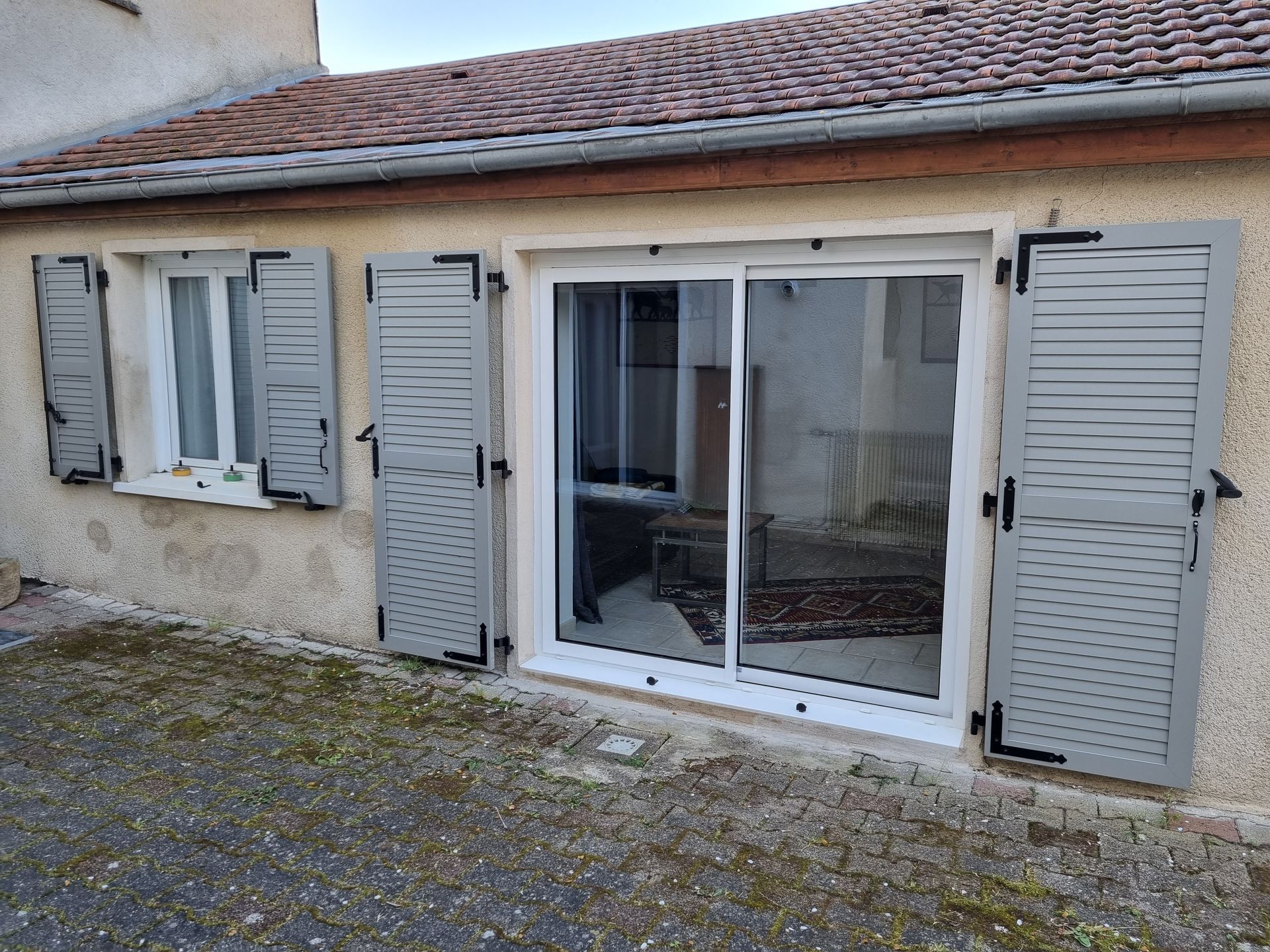 Extérieur d’une maison avec des volets gris clair et une grande baie vitrée coulissante donnant sur une terrasse en gravier.