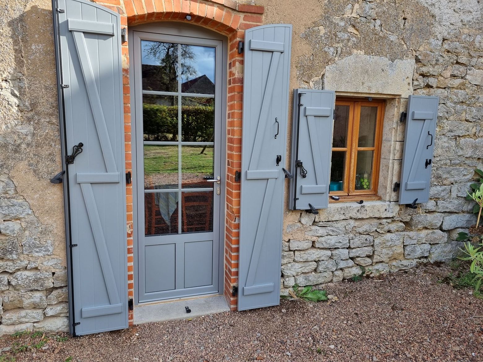 Façade de maison en pierre avec une porte et des volets gris, ainsi qu’une petite fenêtre aux volets bleus.