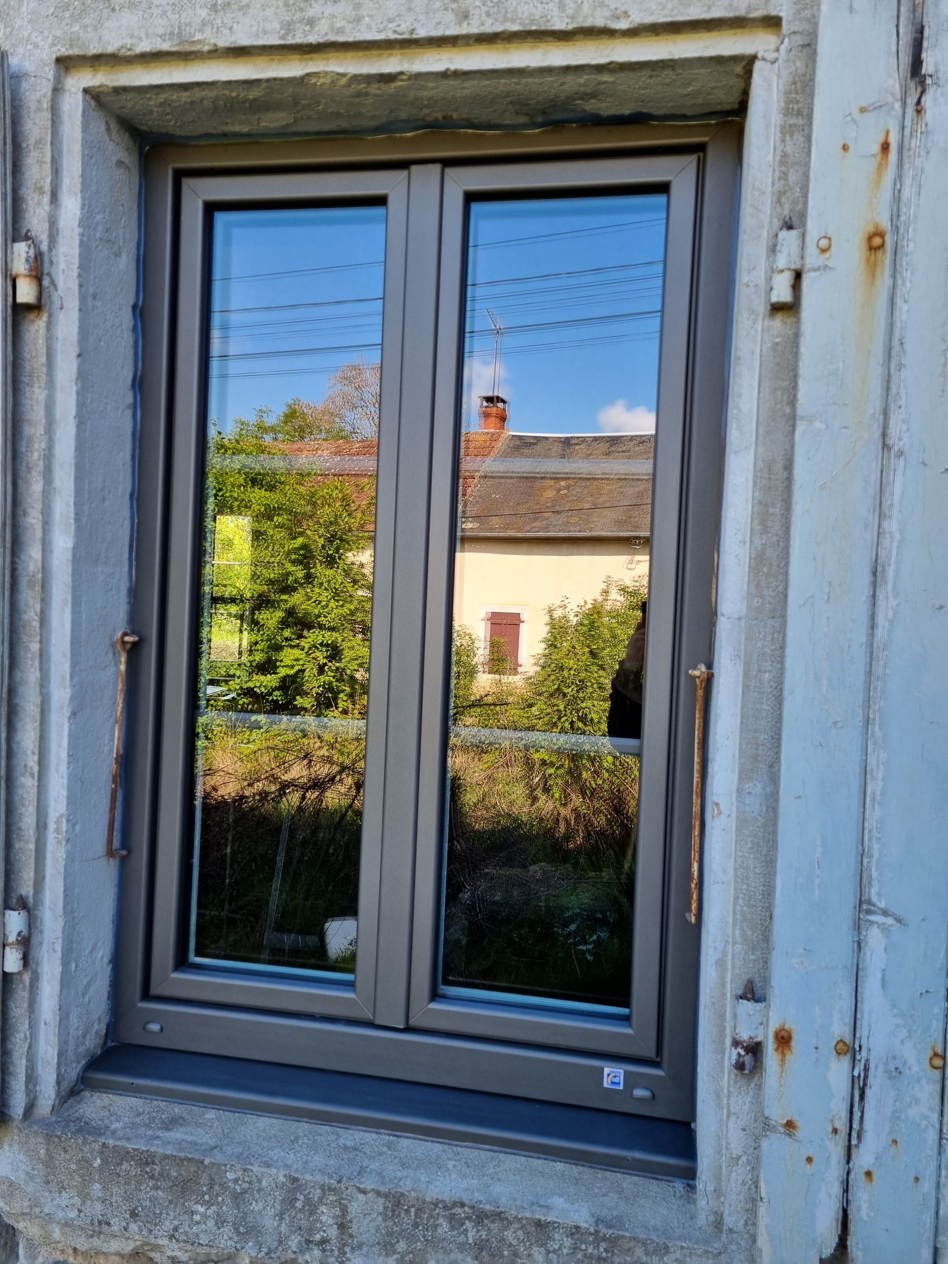 Fenêtre au cadre gris dans un mur de pierre, reflétant une maison et des arbres à l’extérieur.