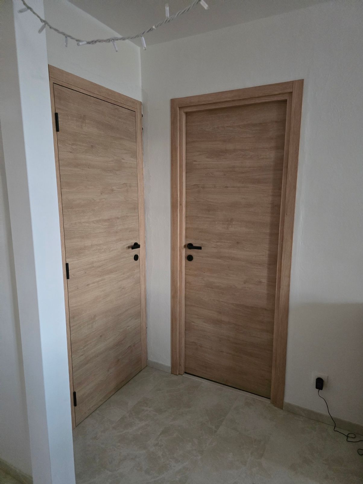 Coin intérieur avec deux portes en bois clair, une pièce peu meublée et un sol carrelé.