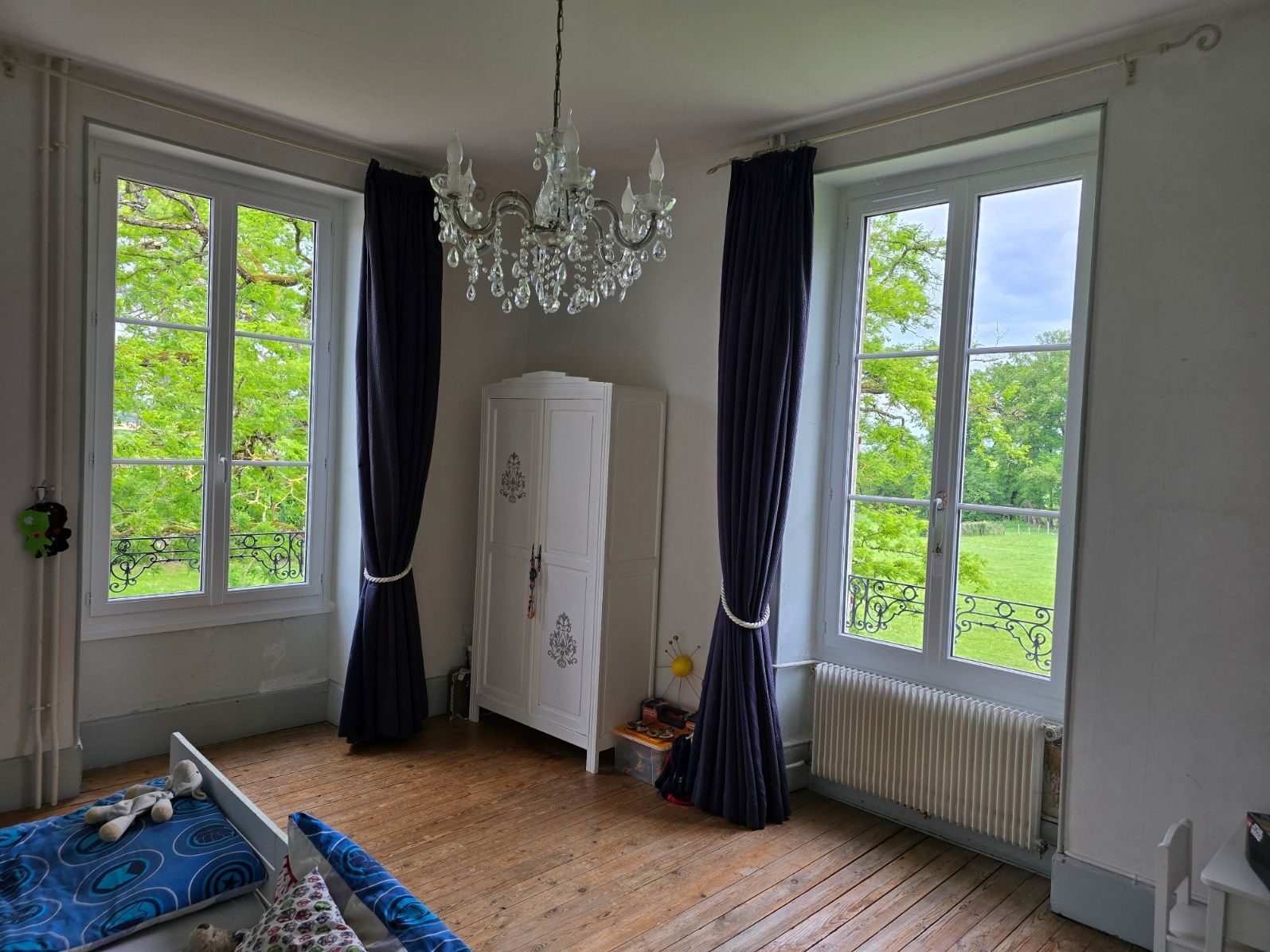 Pièce lumineuse avec de grandes fenêtres, un lustre, des rideaux sombres, une armoire blanche et un canapé bleu.