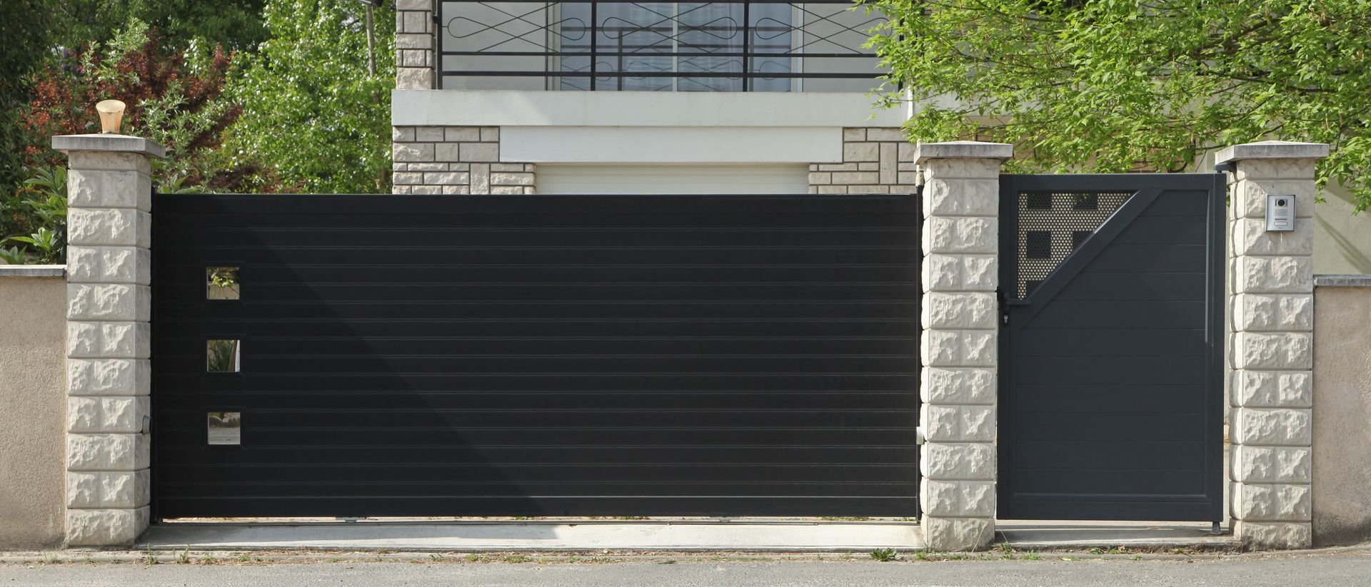 Entrée d’une maison moderne avec un portail coulissant noir et un portillon piéton sur le côté, entre des piliers en pierre.