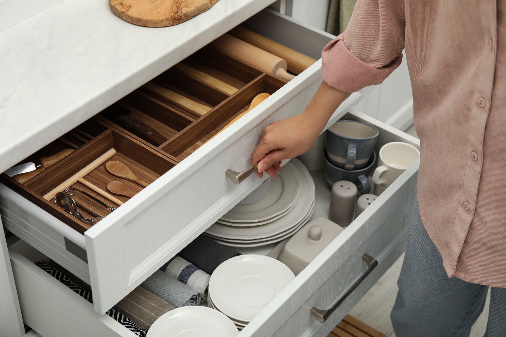 Personne ouvrant un tiroir de cuisine avec des ustensiles et des assiettes empilées à côté d’un évier.