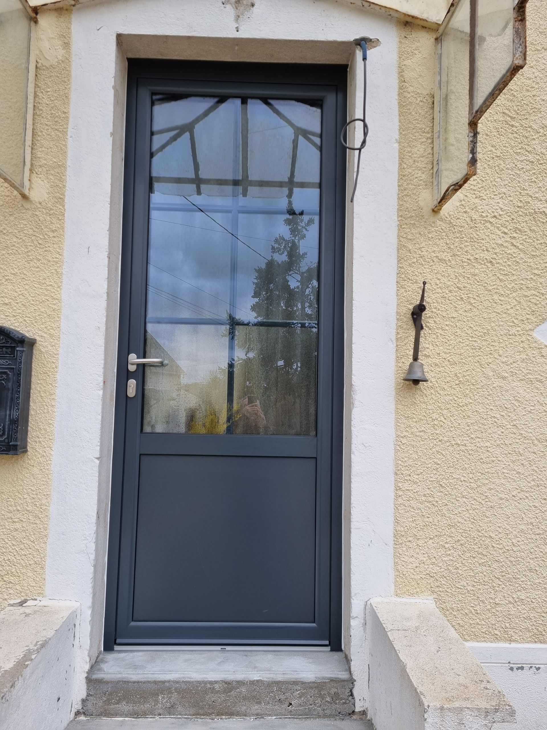 Porte d’entrée grise avec panneau vitré dans une entrée en stuc beige aux encadrements blancs.