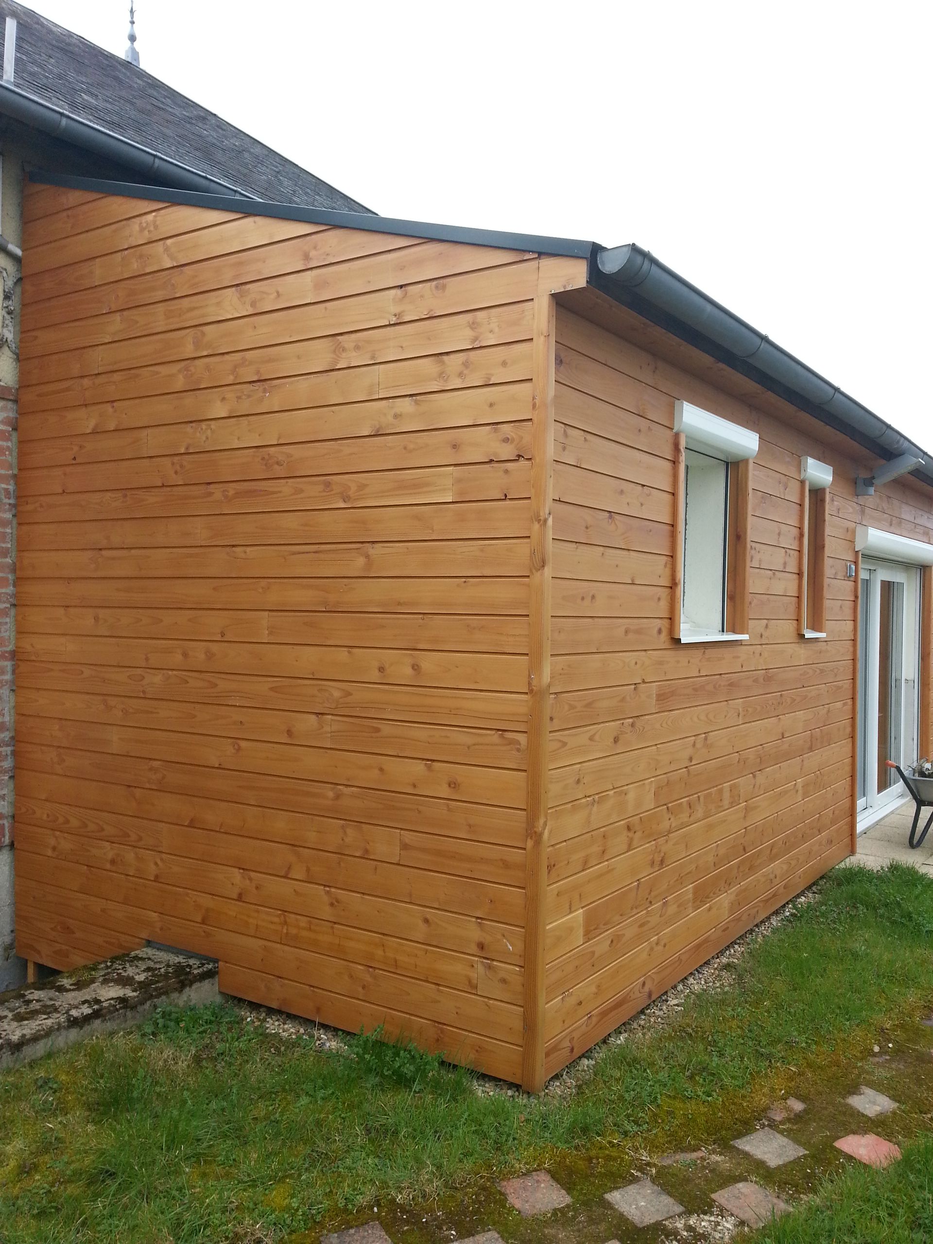 Façade latérale en bois d’une maison, avec une petite fenêtre à encadrement blanc et de l’herbe.
