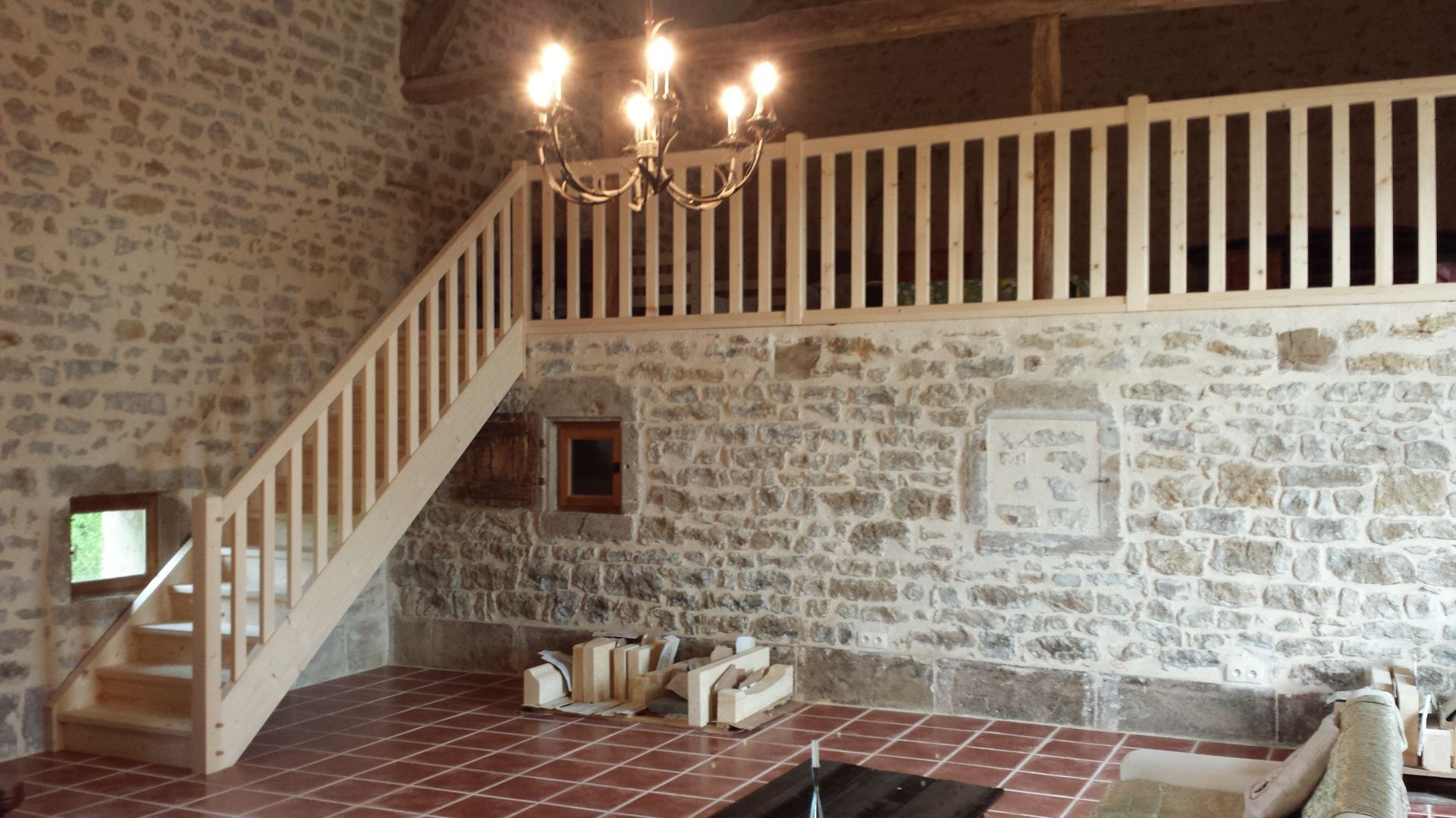 Pièce aux murs de pierre, avec un sol en carreaux rouges, un escalier blanc menant à une mezzanine et un lustre suspendu.