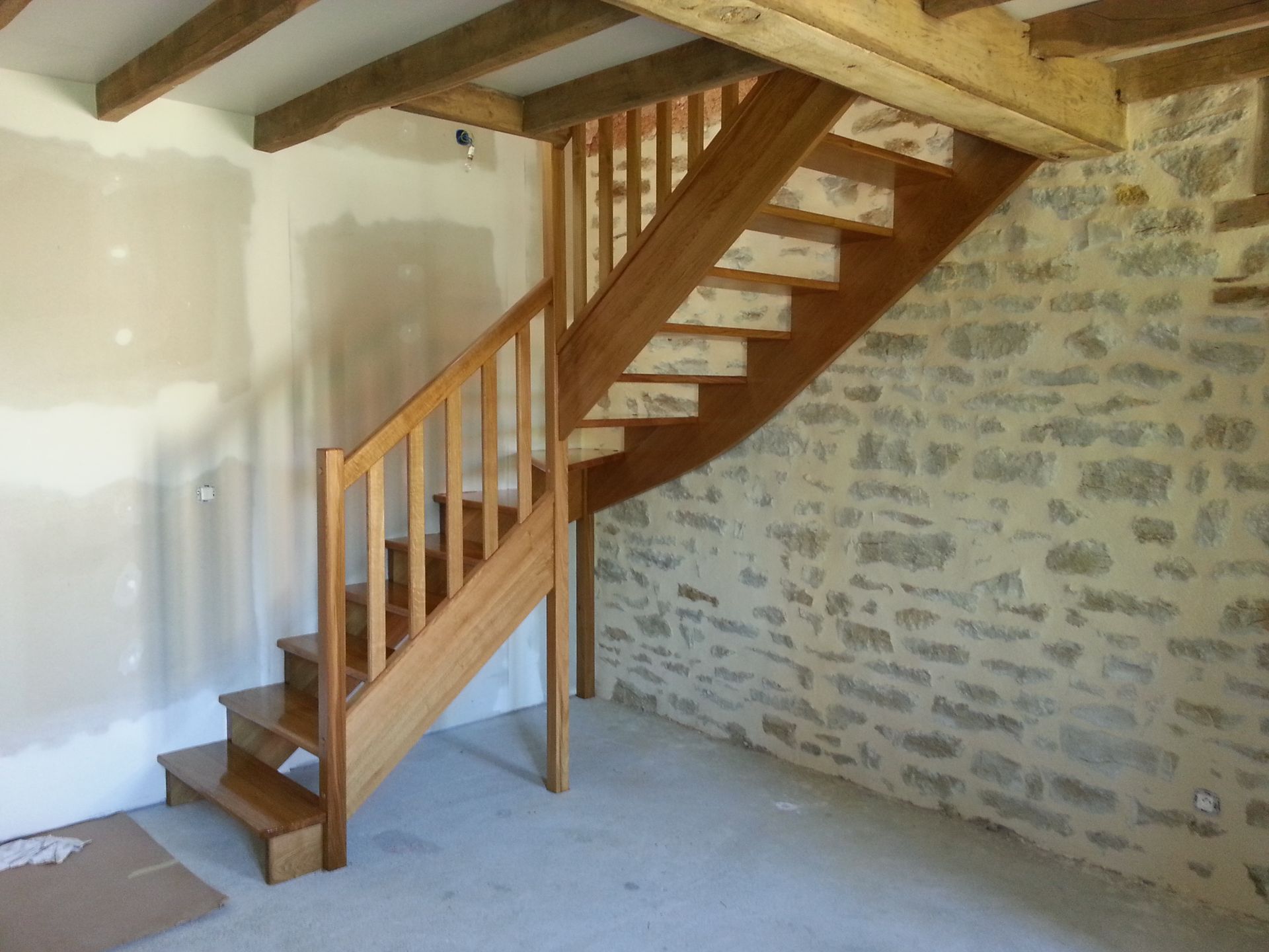Escalier en bois dans une pièce rustique en pierre, avec des poutres apparentes et des murs enduits.