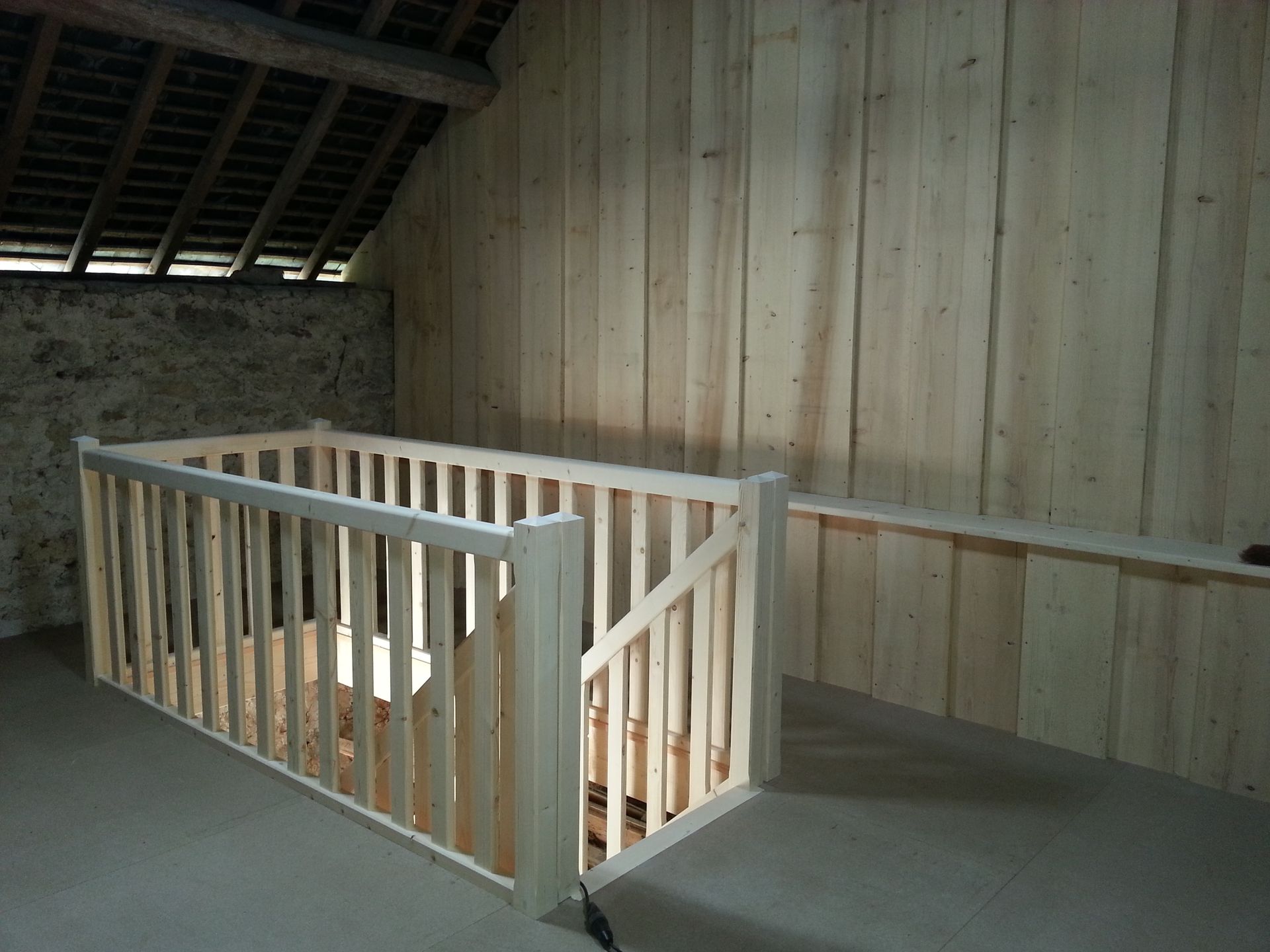 Cage d’escalier en bois avec des rampes blanches, dans un intérieur inachevé aux allures de grenier.