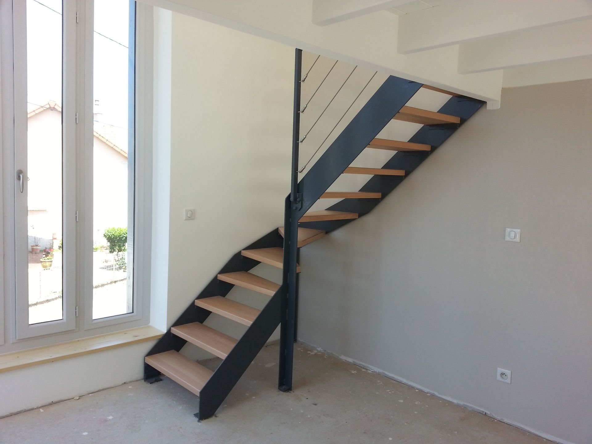 Escalier intérieur en construction, bordé de hautes fenêtres, avec des marches inachevées et des poteaux de soutien noirs.