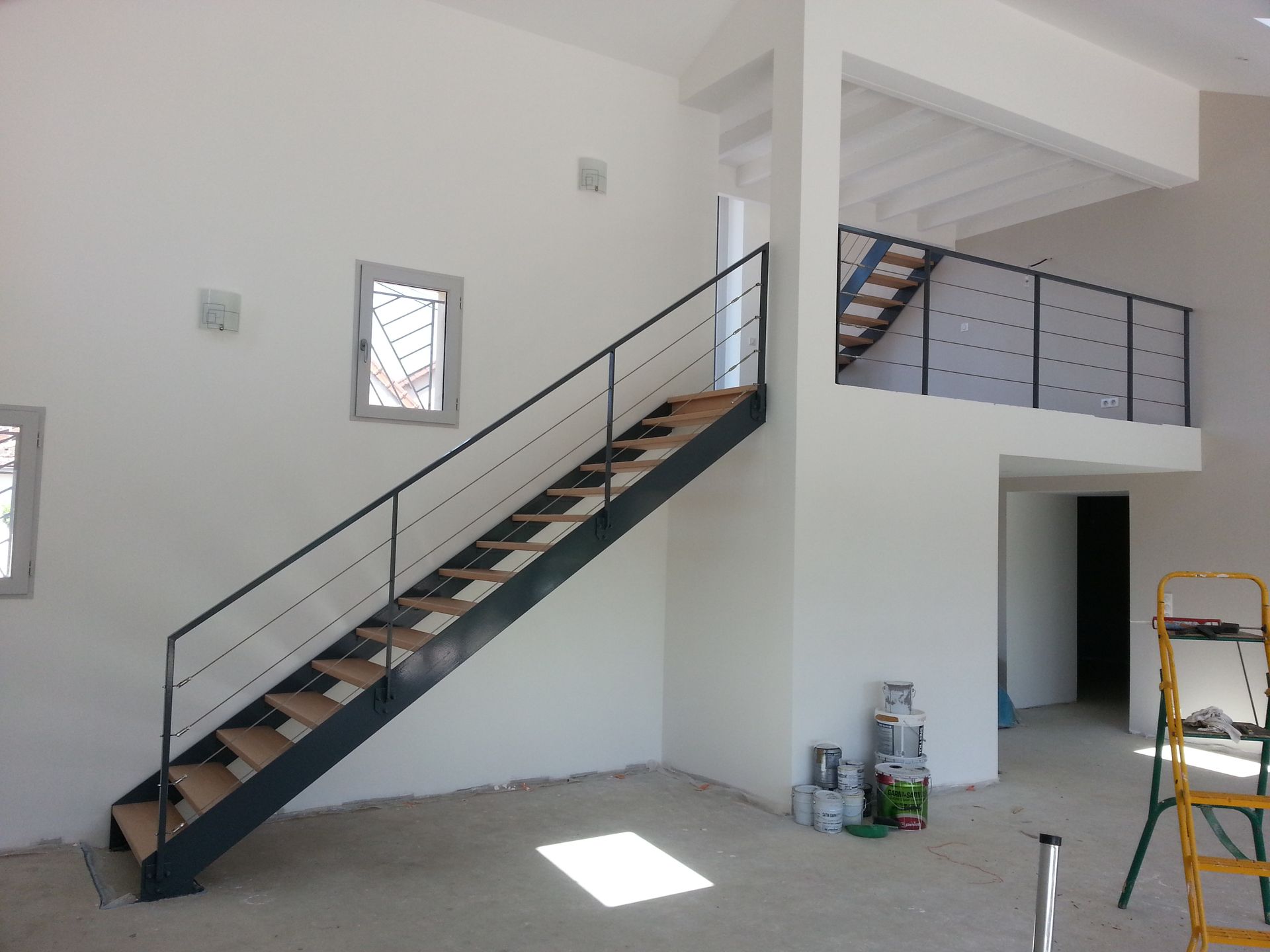 Pièce intérieure avec un escalier en métal noir, des murs blancs et une rambarde à l’étage, dans un espace lumineux non aménagé.