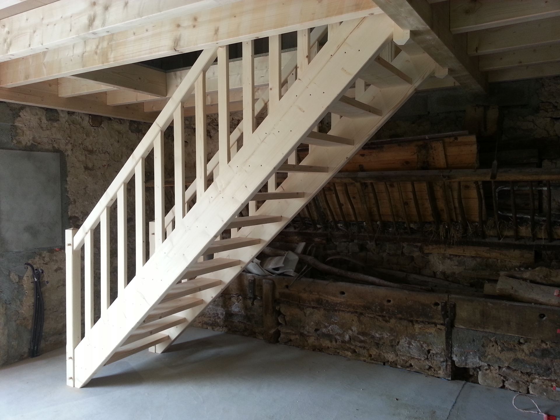 Escalier en bois blanc sous une mezzanine, dans un intérieur inachevé aux murs en brique apparente et en béton.