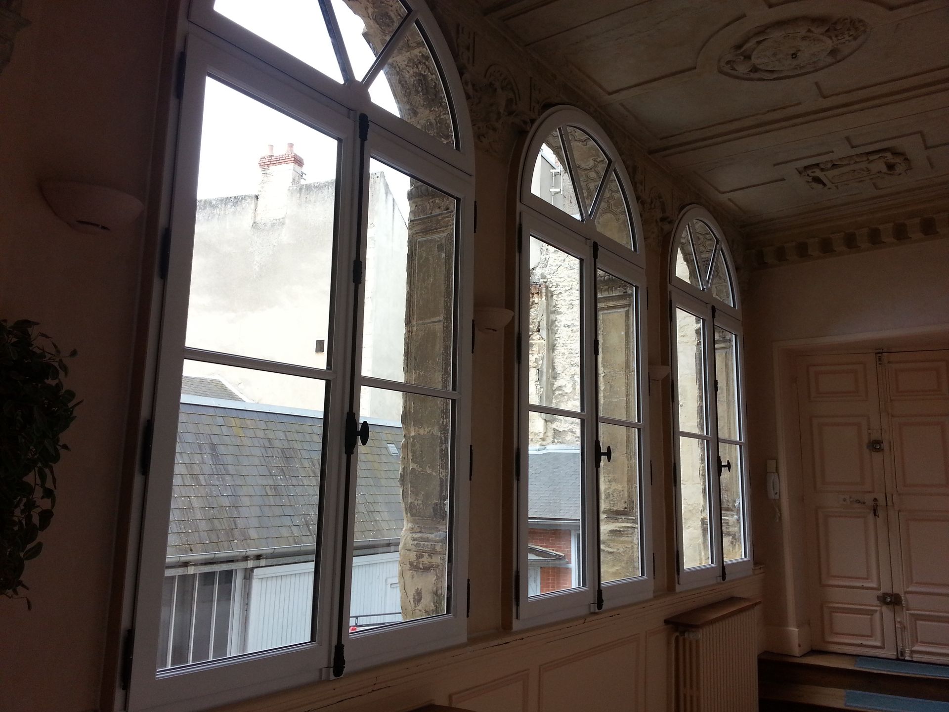 Intérieur baigné de soleil avec de hautes fenêtres en arc donnant sur des toits et des balcons.