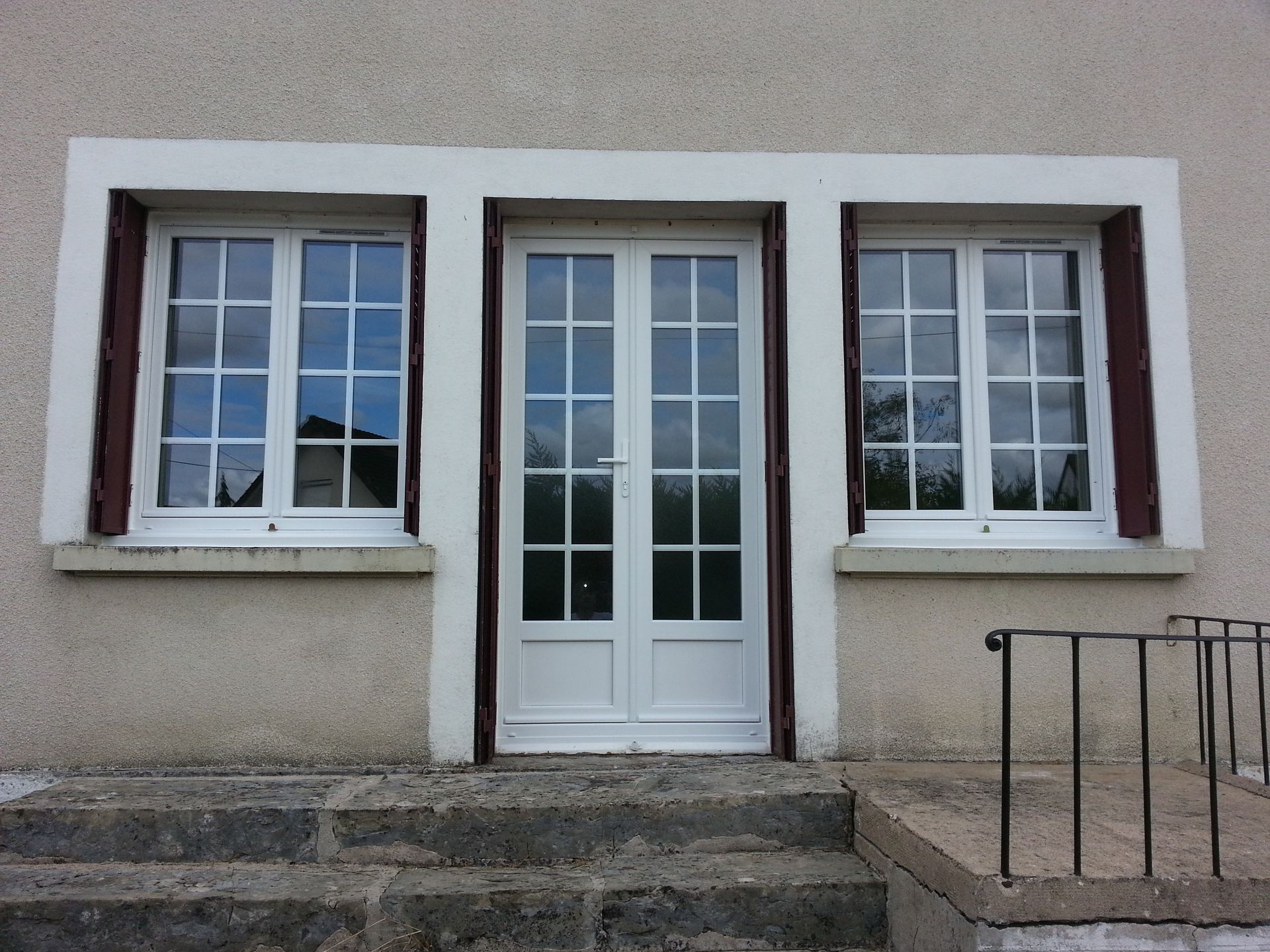Entrée d’un bâtiment avec une double porte blanche, deux fenêtres, des marches en pierre et une rampe métallique.