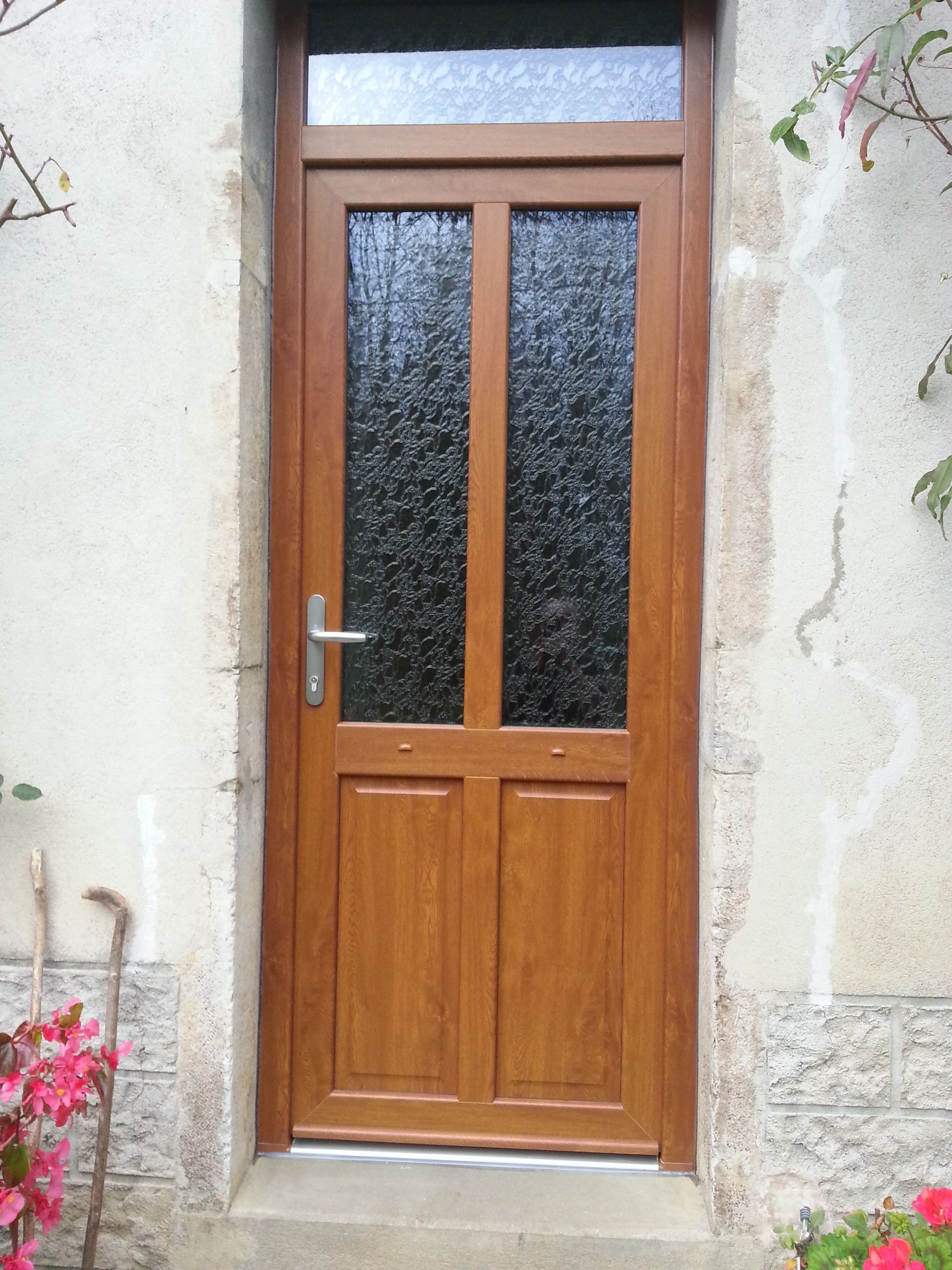 Porte d’entrée en bois avec des panneaux de verre dépoli, encastrée dans un mur de pierre, et des fleurs en bas à gauche.