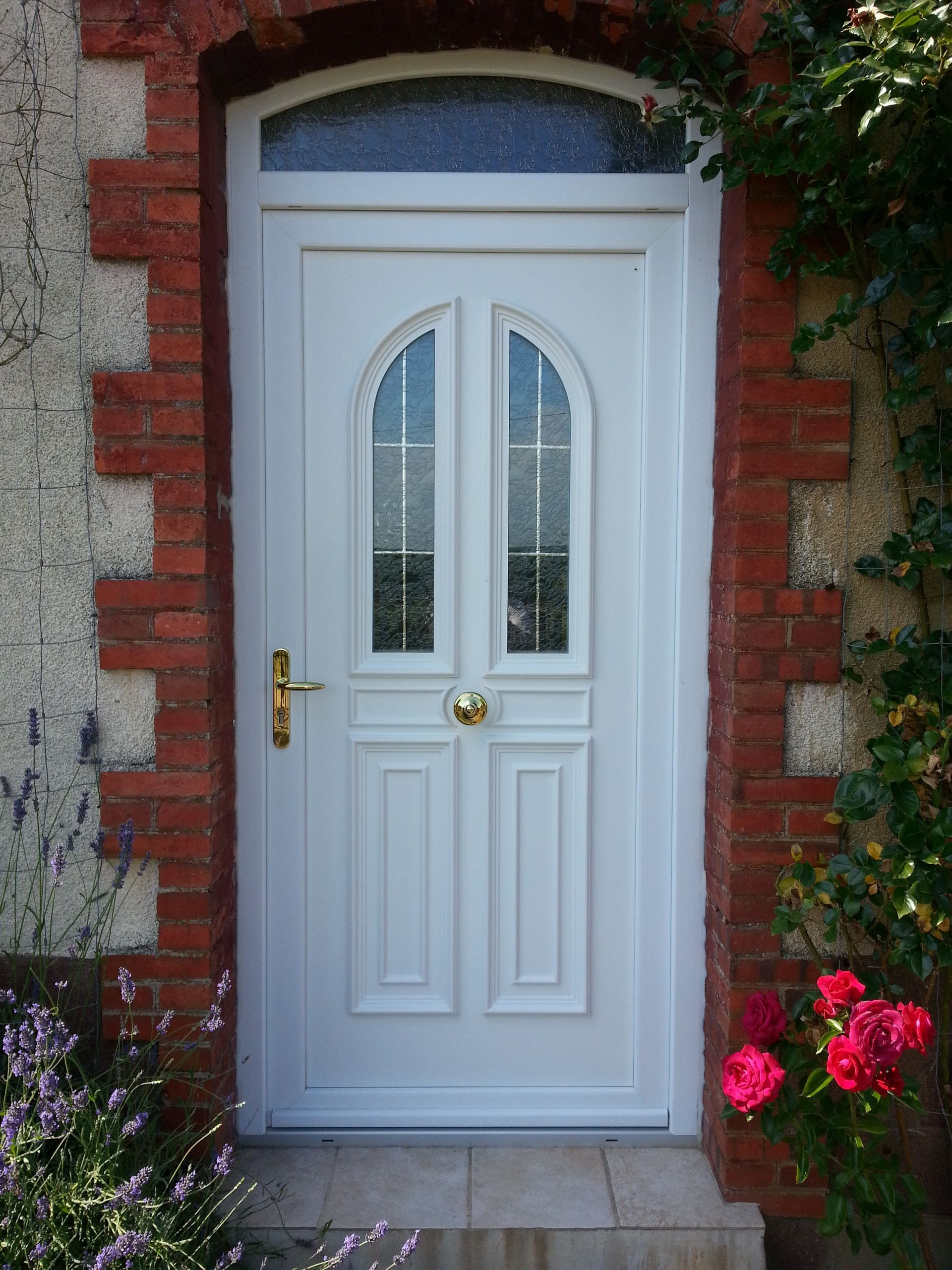 Porte d’entrée blanche avec des vitraux en arc, une poignée en laiton et des fleurs de chaque côté, dans une entrée en briques.