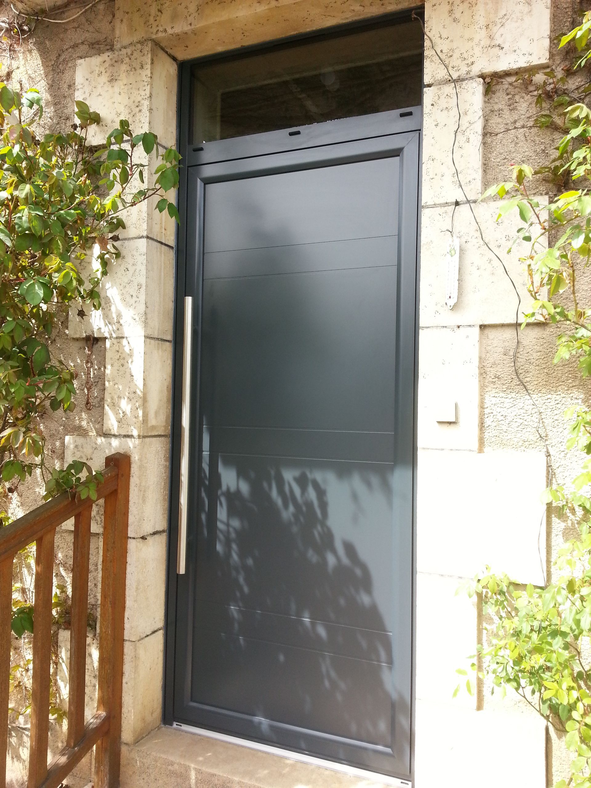Porte en métal gris foncé encastrée dans un mur de pierre, avec de la verdure de chaque côté.