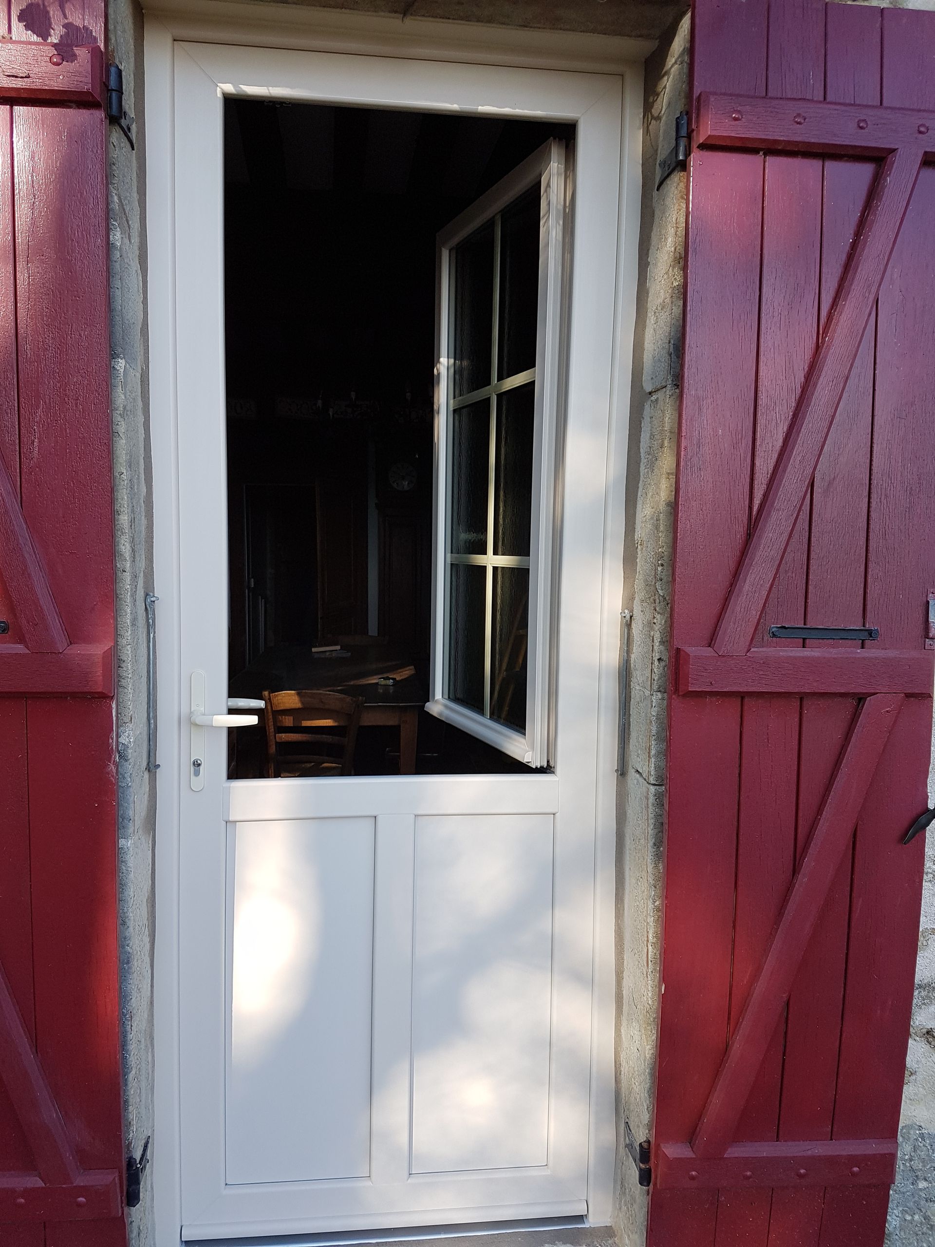 Porte blanche avec une fenêtre ouverte dans un mur rouge en bois, donnant sur un intérieur sombre.