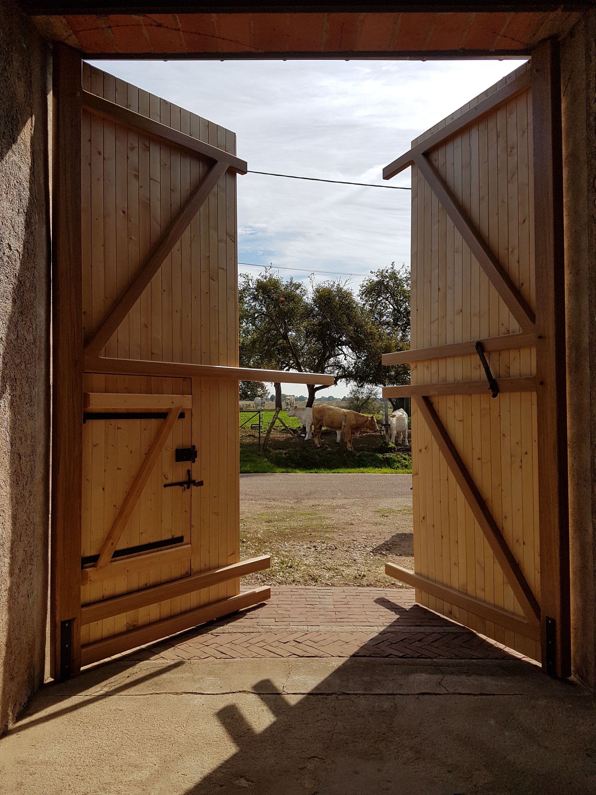 Portails en bois ouverts encadrant une cour ensoleillée, avec des arbres et du gravier au-delà.