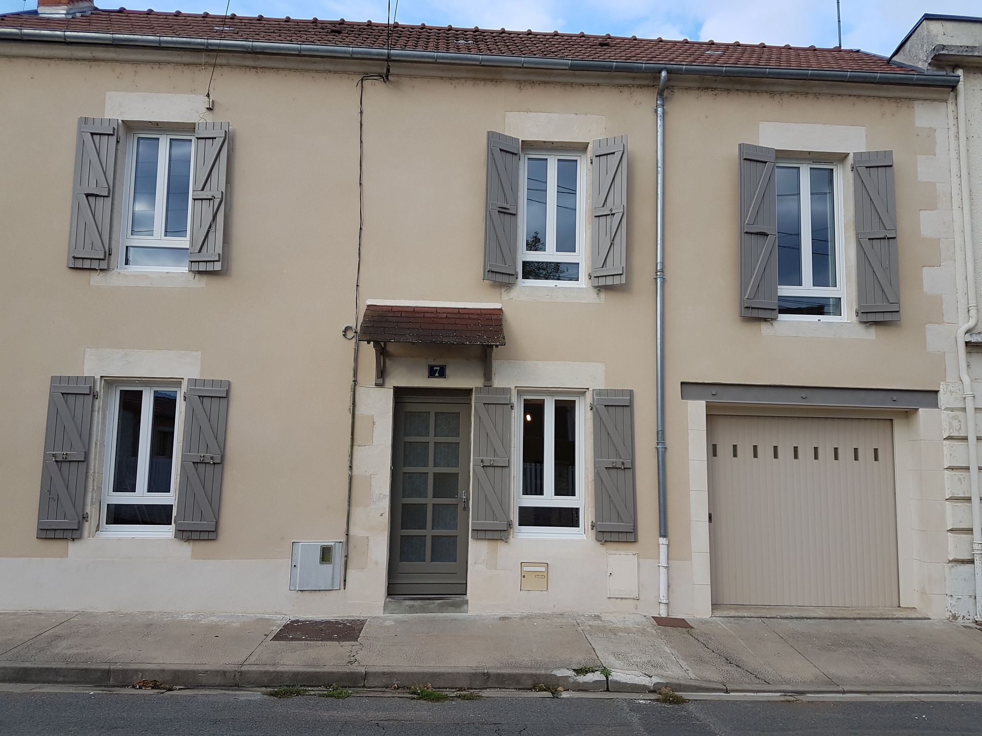 Une maison beige à deux étages, avec des volets, une porte centrale et un garage à droite.