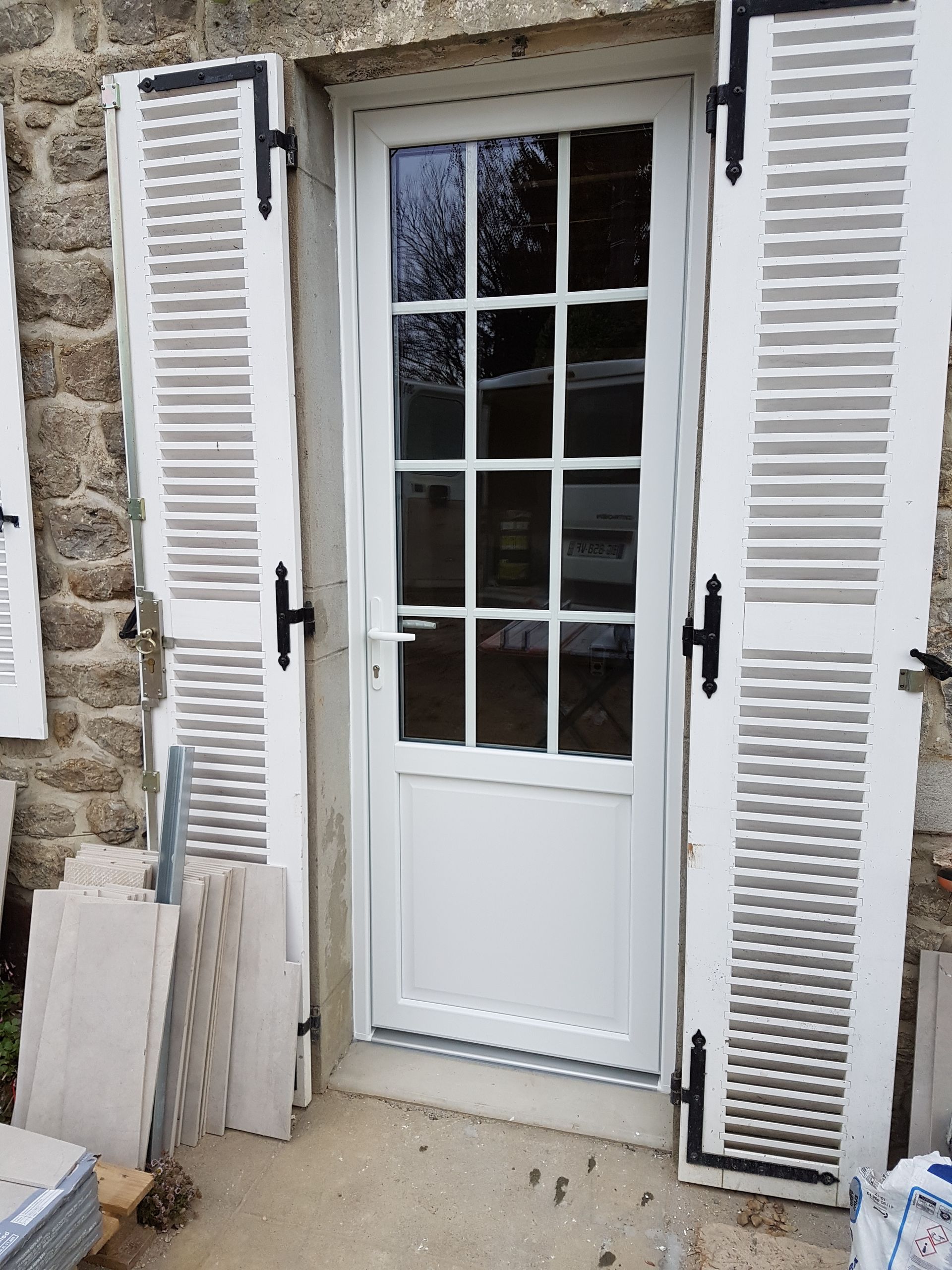 Porte blanche à vitres, flanquée de volets blancs ouverts, sur la façade en pierre d’une maison.