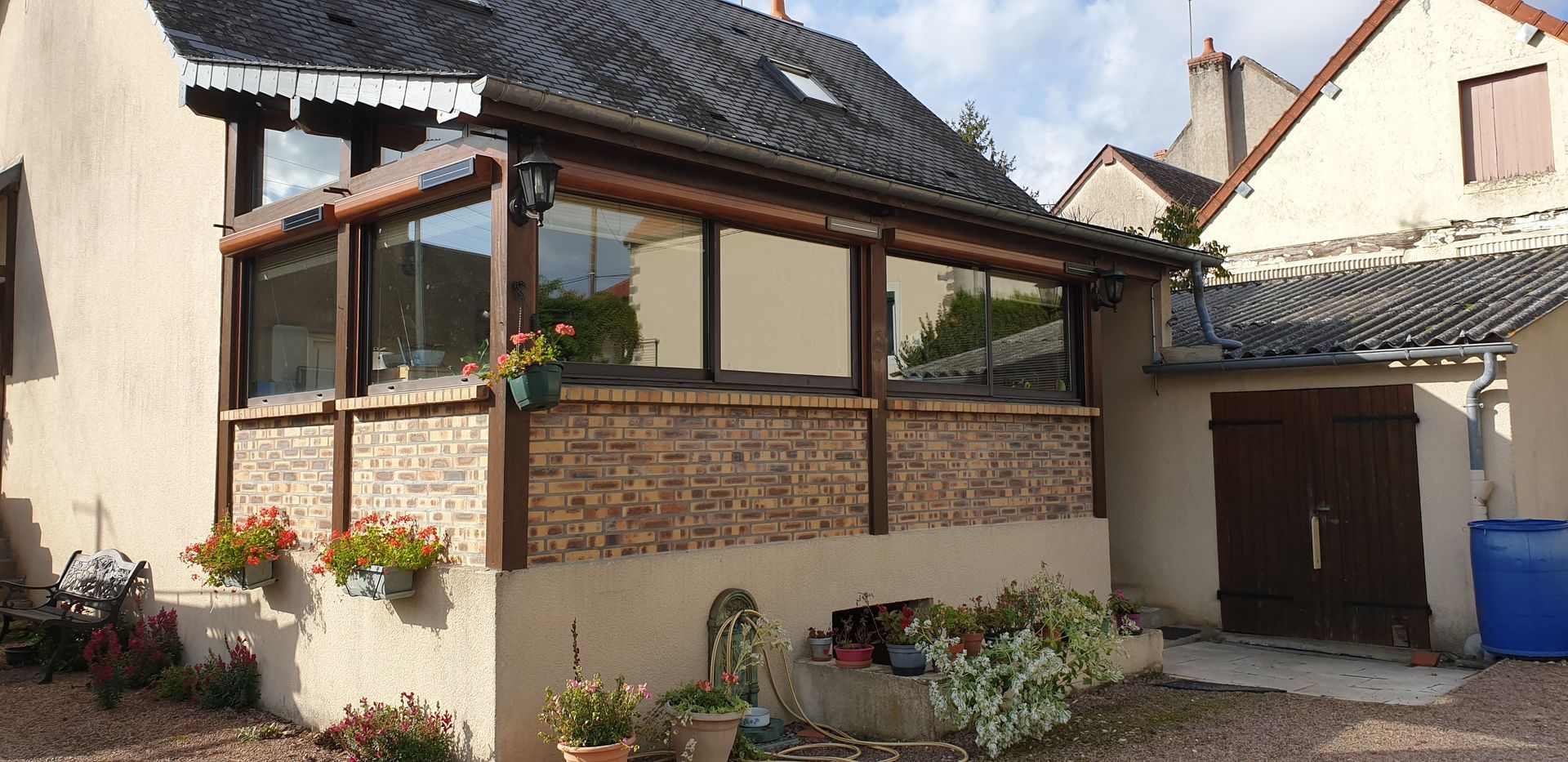 Cour intérieure d’une petite maison en pierre, avec des fleurs en pot, une véranda en bois et un tonneau bleu à côté d’une porte de garage.