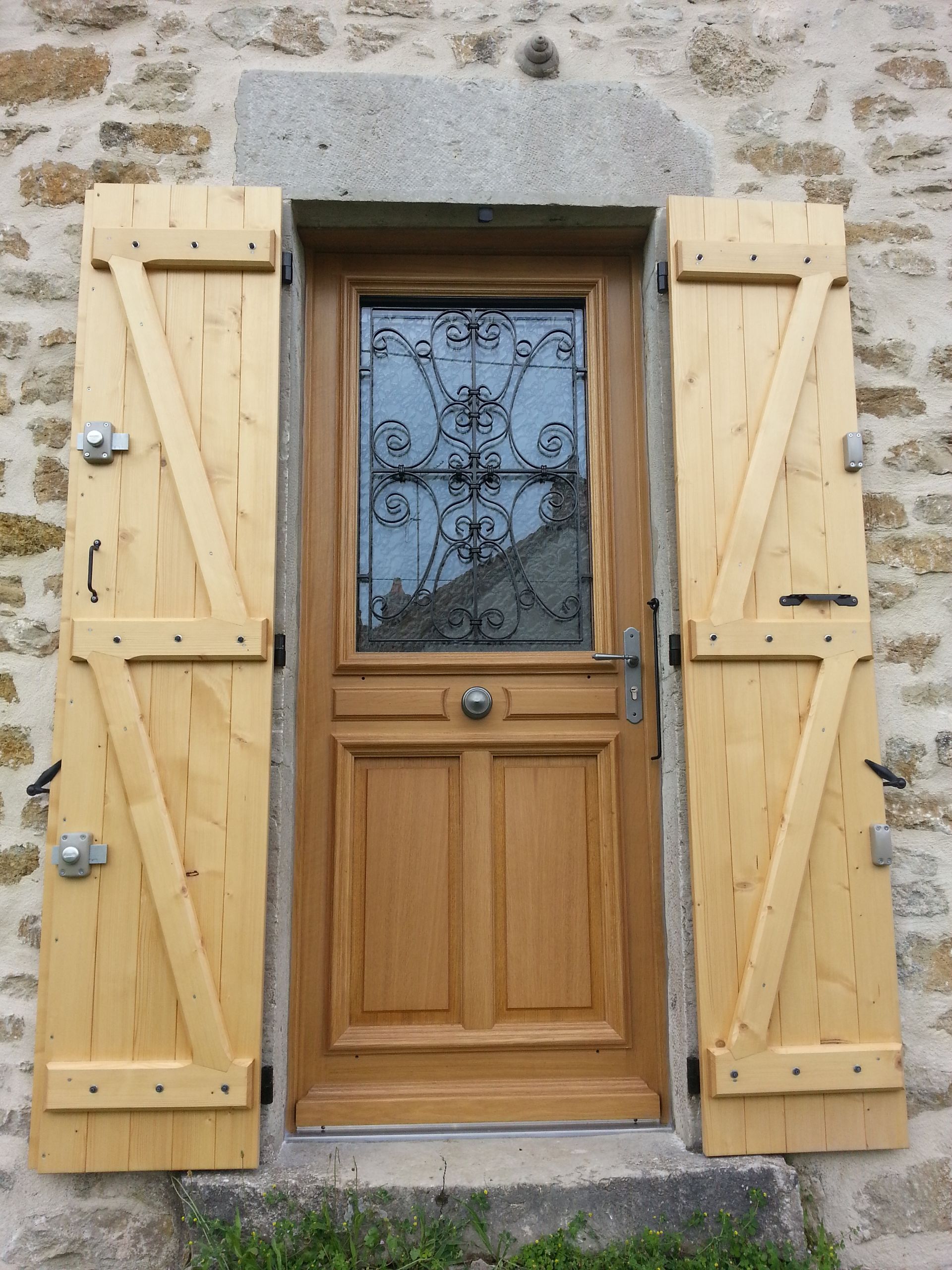 Porte d’entrée en bois avec vitrage décoratif, encadrée de volets en bois clair sur un mur en pierre.