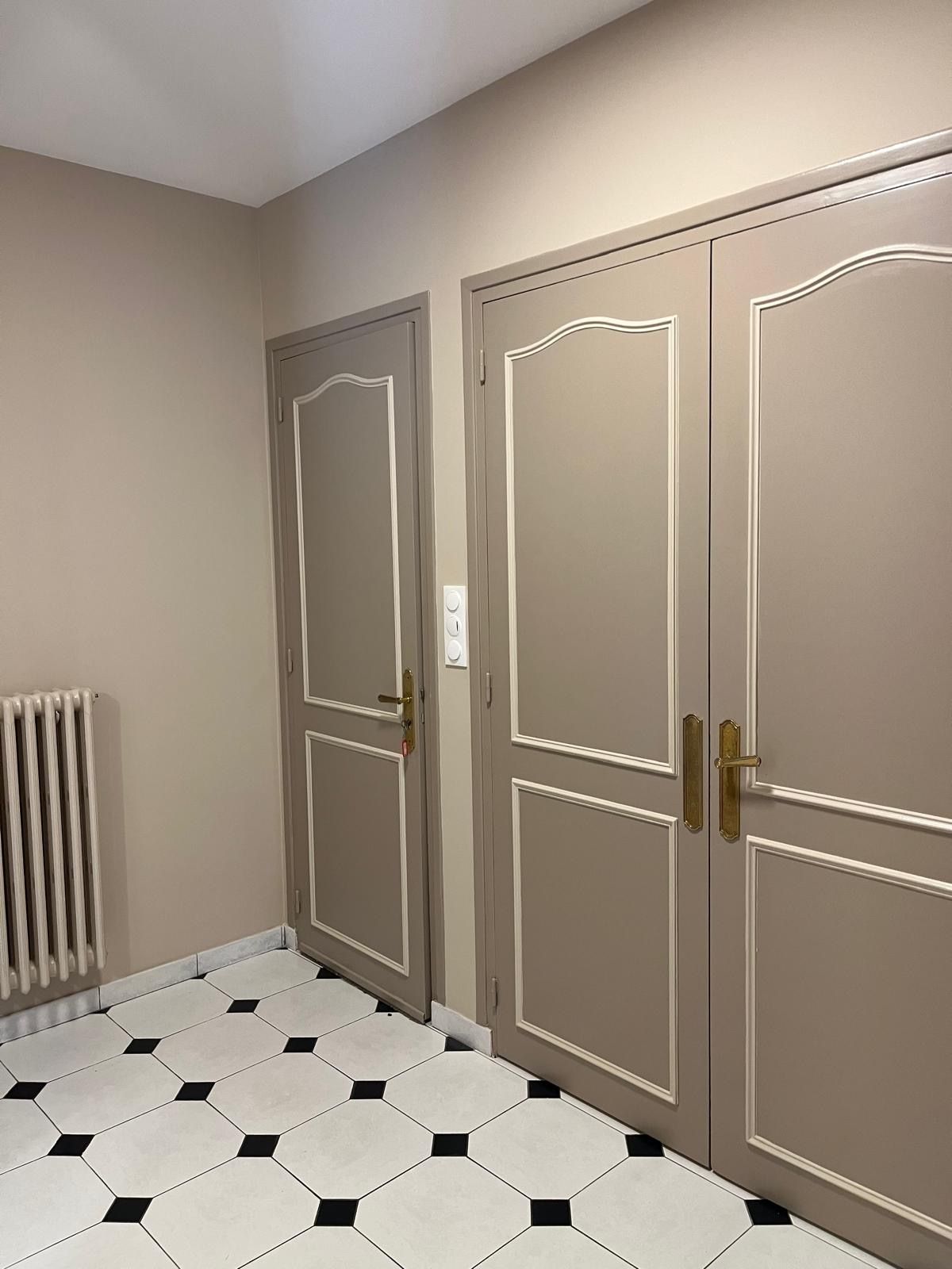 Une pièce aux tons neutres présente deux portes taupe avec des moulures blanches et des poignées dorées, à côté d'un radiateur sur un sol carrelé.