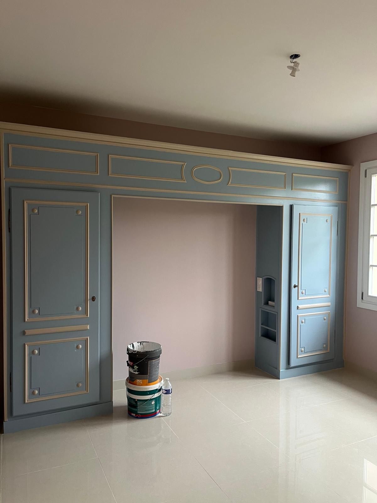 Un meuble de rangement sur mesure gris pour chambre à coucher, avec des panneaux à bordures dorées et une alcôve encastrée, flanqué de hautes armoires et d'une fenêtre.