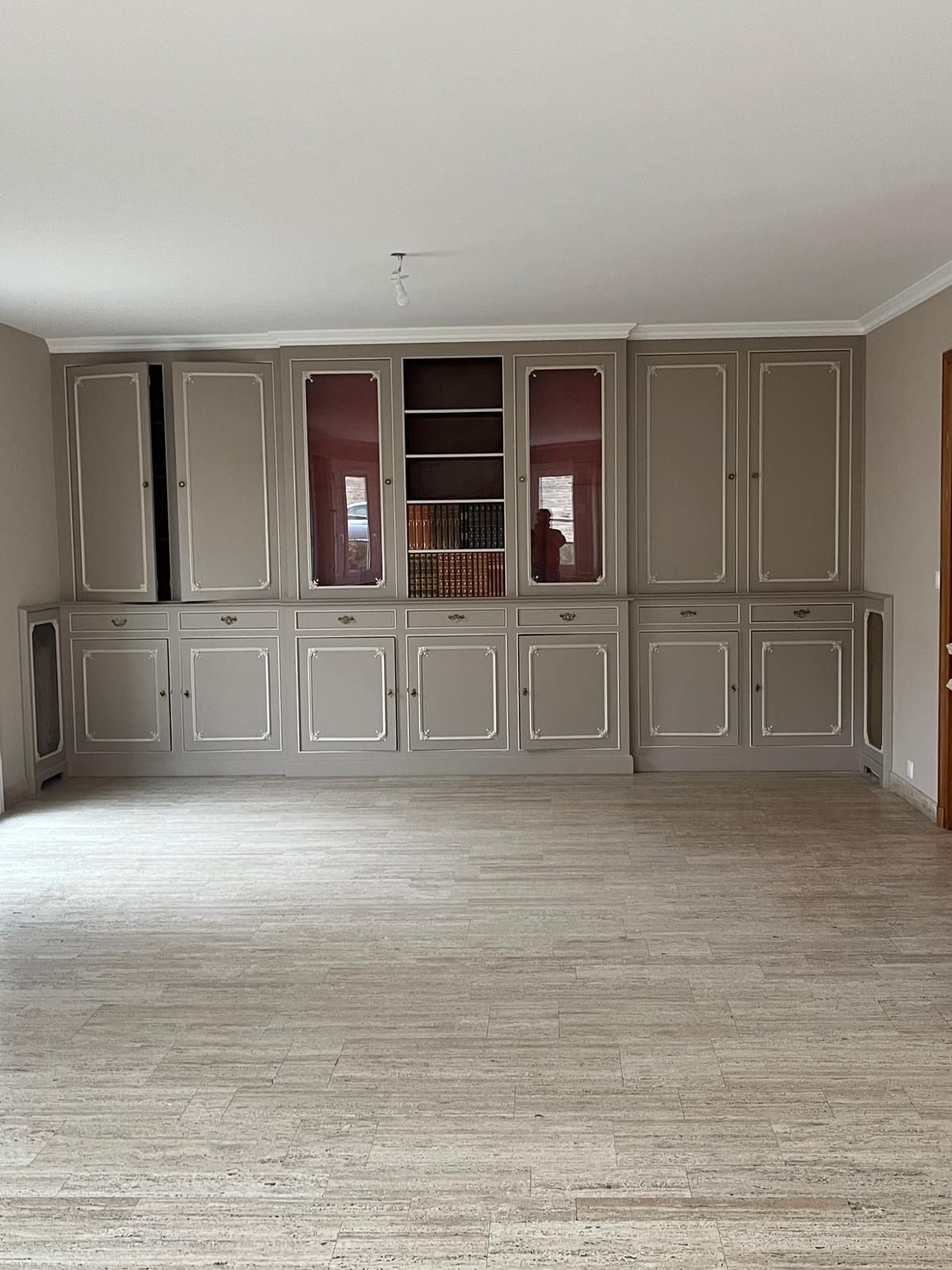 Un large meuble mural sur mesure aux tons neutres, avec vitrines, étagères et placards, adossé à un sol carrelé en pierre.