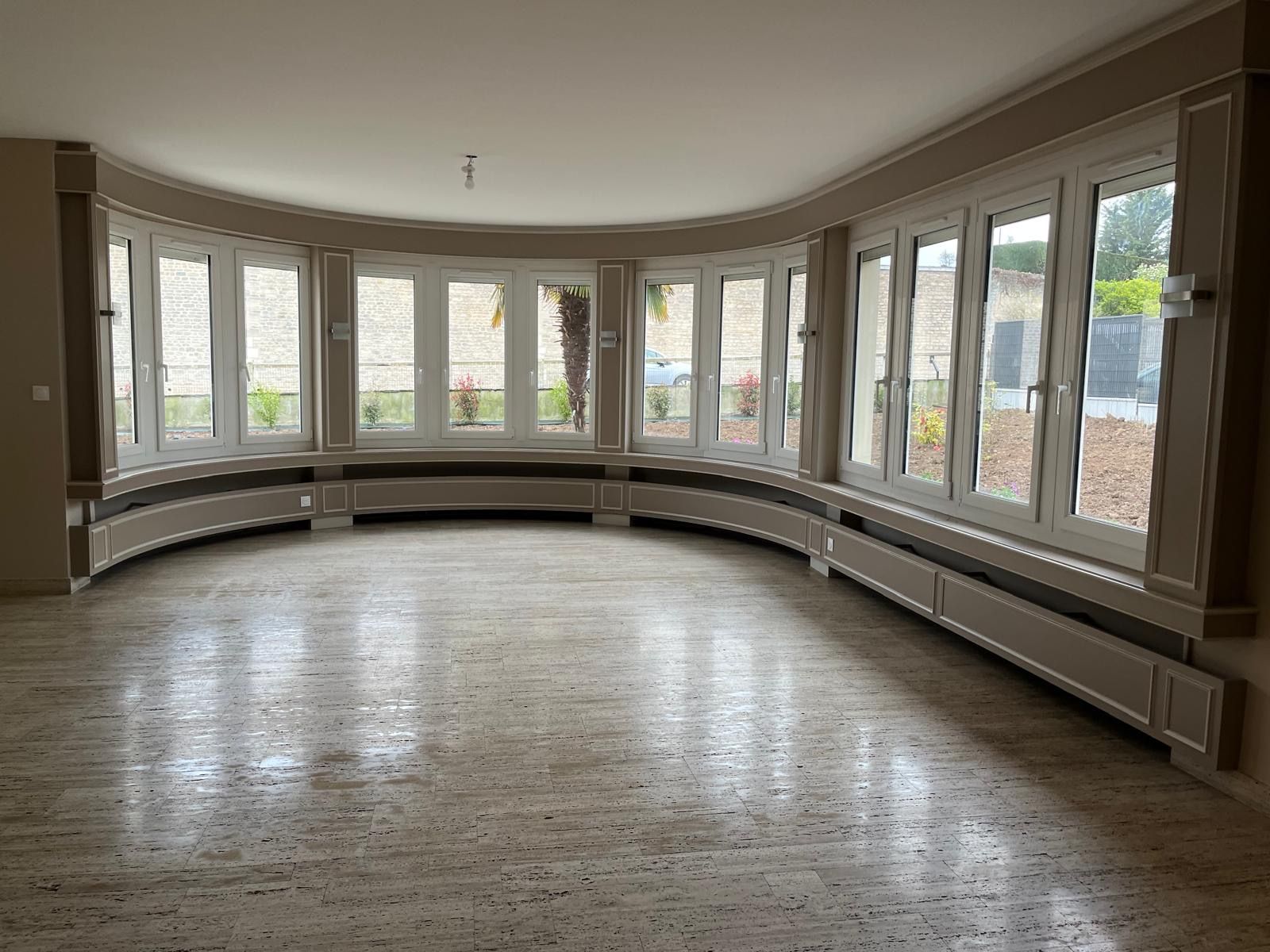 Une pièce spacieuse en demi-cercle avec de grandes fenêtres, un sol en pierre beige et des cache-radiateurs incurvés le long de la base des murs.