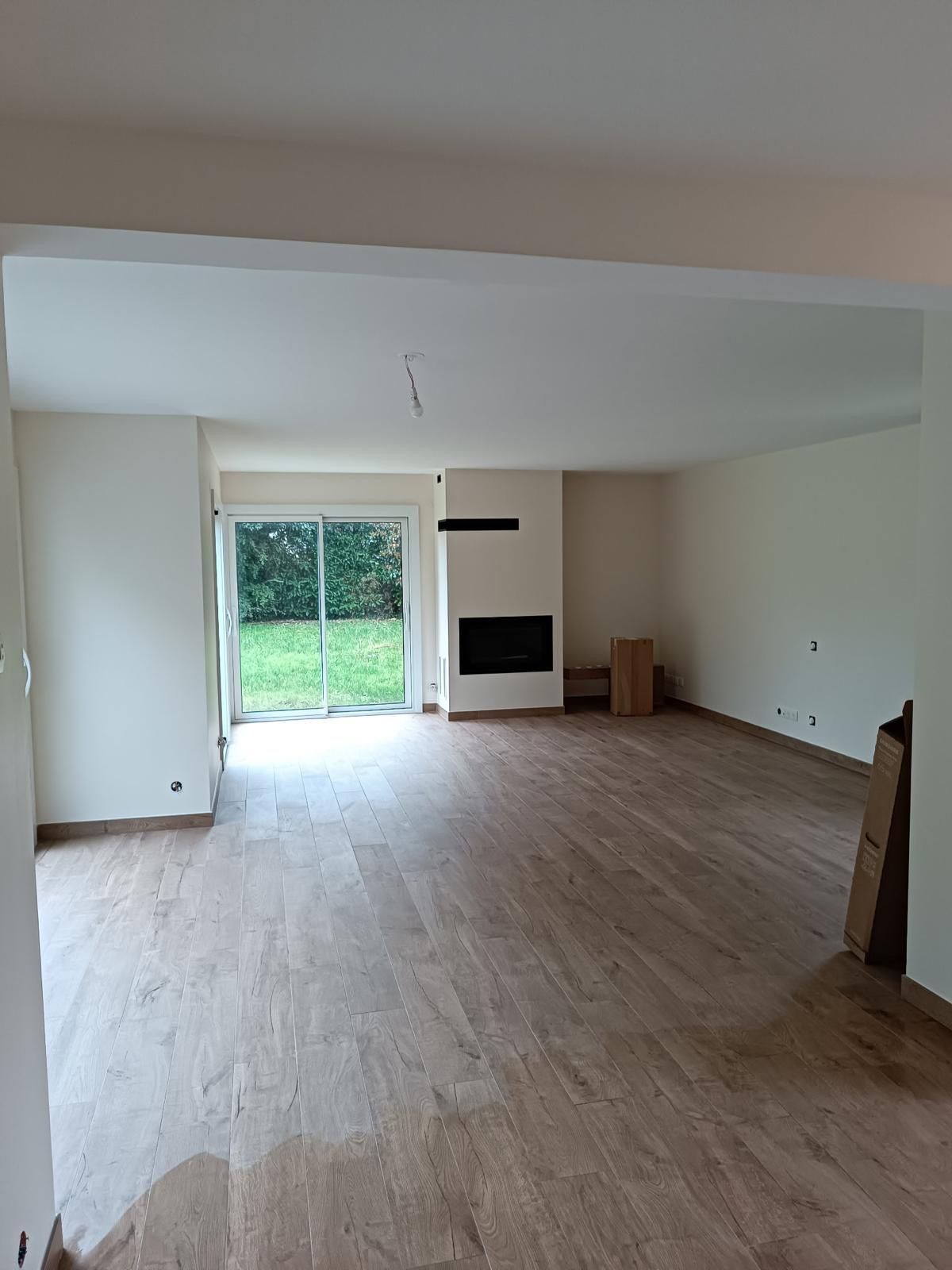 Un salon vide avec parquet clair, murs couleur crème, une porte-fenêtre coulissante donnant sur une cour et une cheminée.