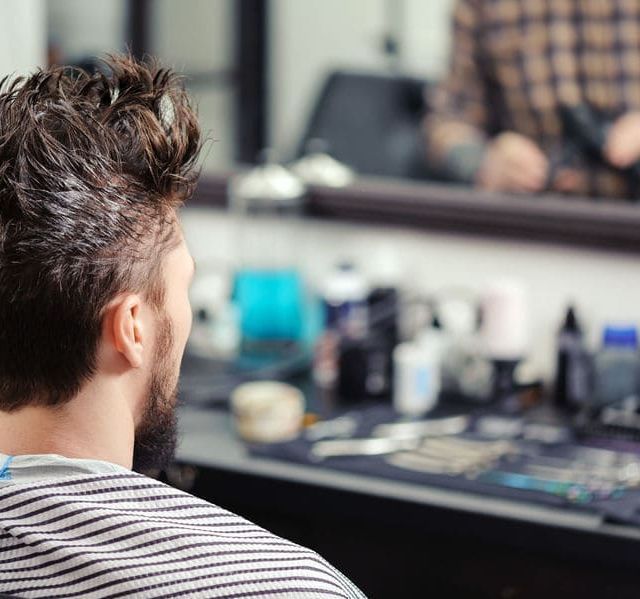 Un hombre con barba está sentado frente a un espejo en una barbería.